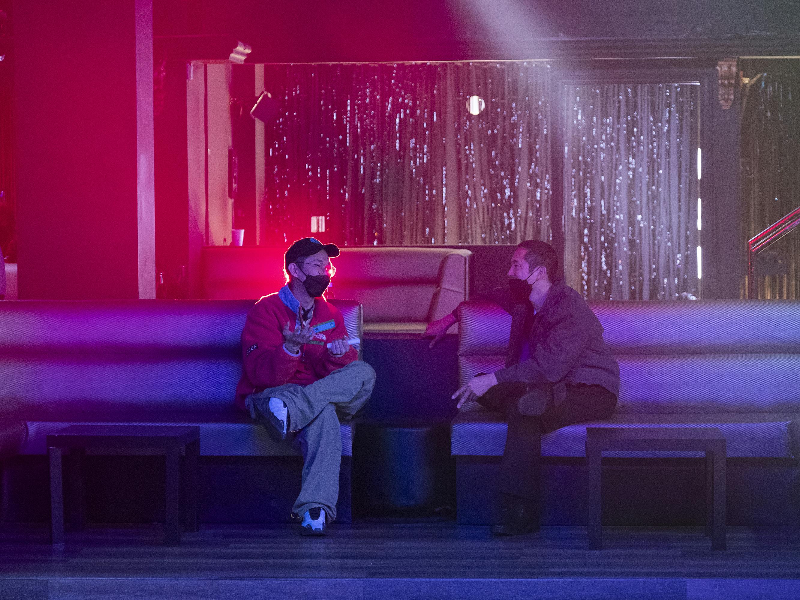 Lee Sung Jin and Steven Yeun sit together in a red-and-purple lit club set. 