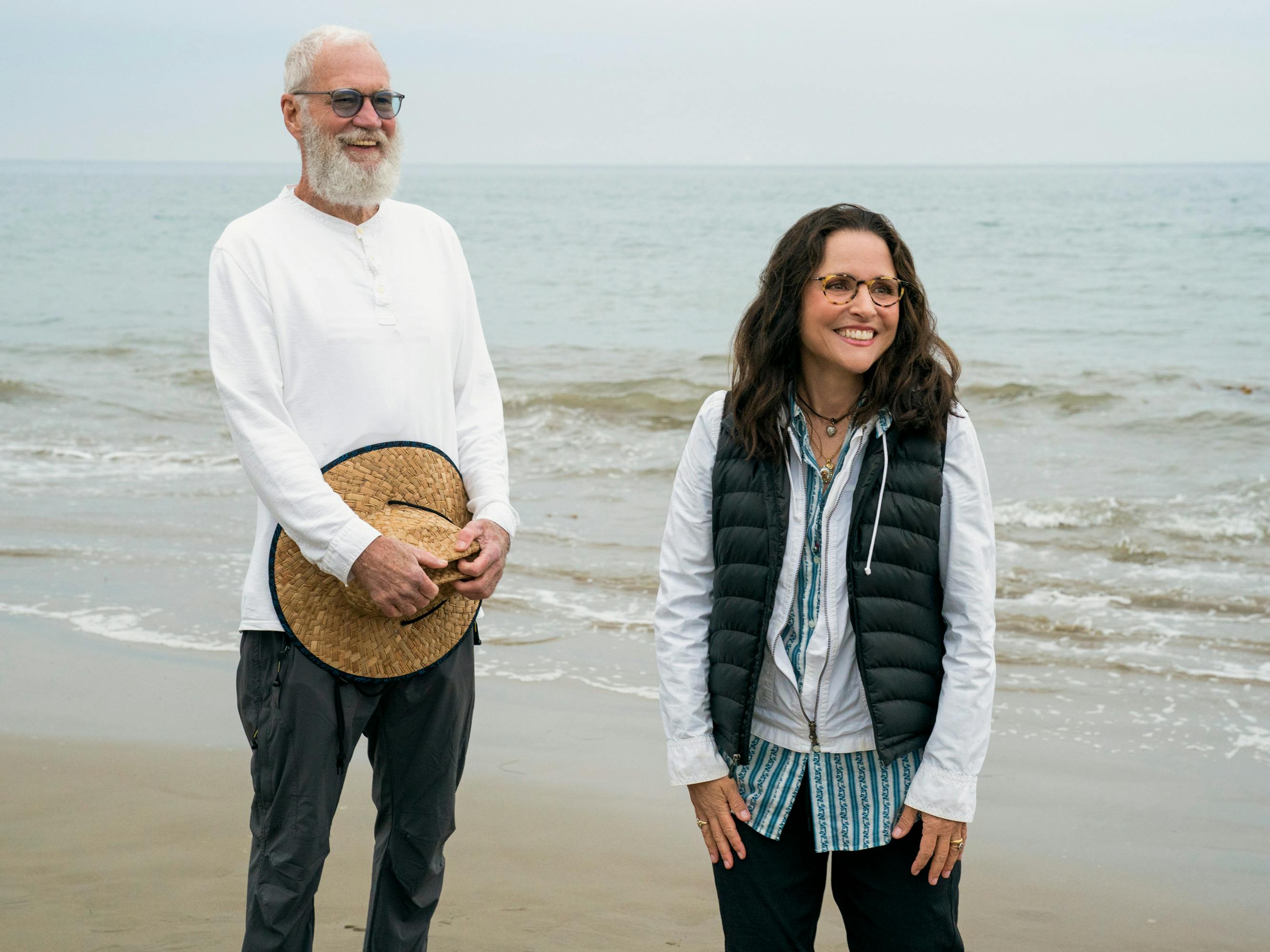 David Letterman and Julia Louis-Dreyfus stand on a beach smiling. Letterman wears a white shirt, dark pants, and a tan hat. Julia wears  a puffer vest, sweatshirt, and glasses.