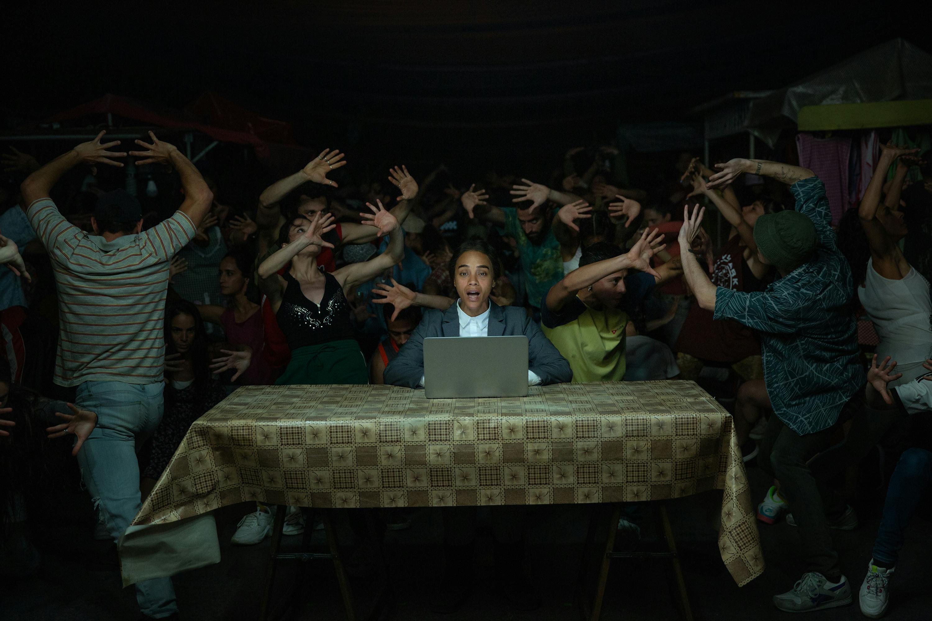 Rita (Zoe Saldaña) sits at a table behind a laptop, as people behind her dance wildly.