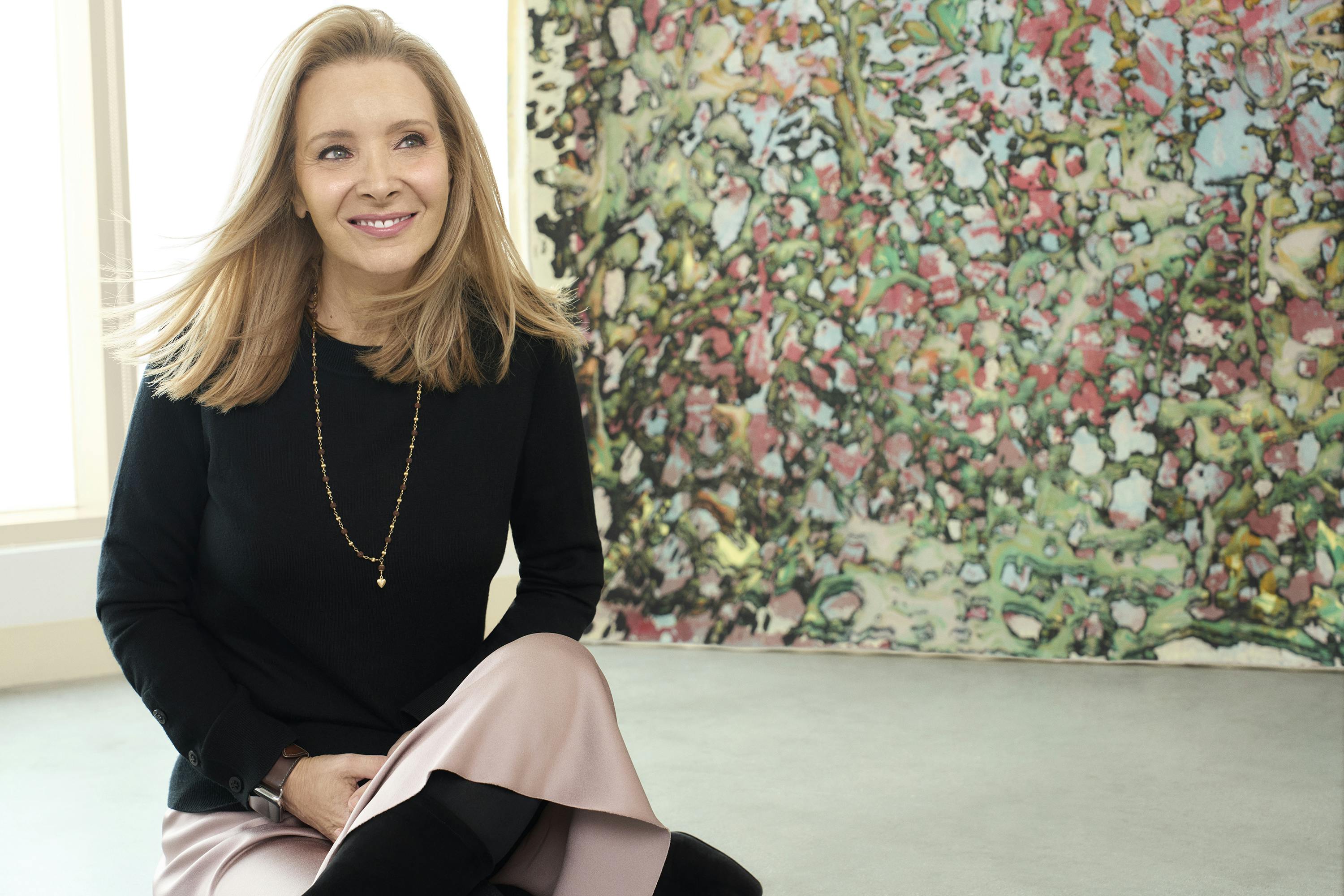 Lisa Kudrow  wears a black ensemble and sits in what looks like an art gallery.