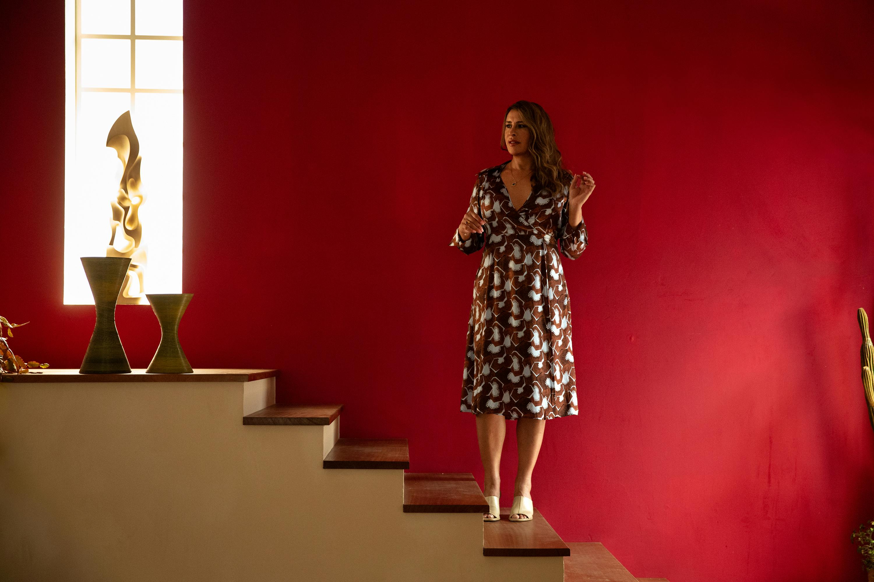 Emilia Pérez (Karla Sofía Gascón) stands on a yellow staircase against a red wall.