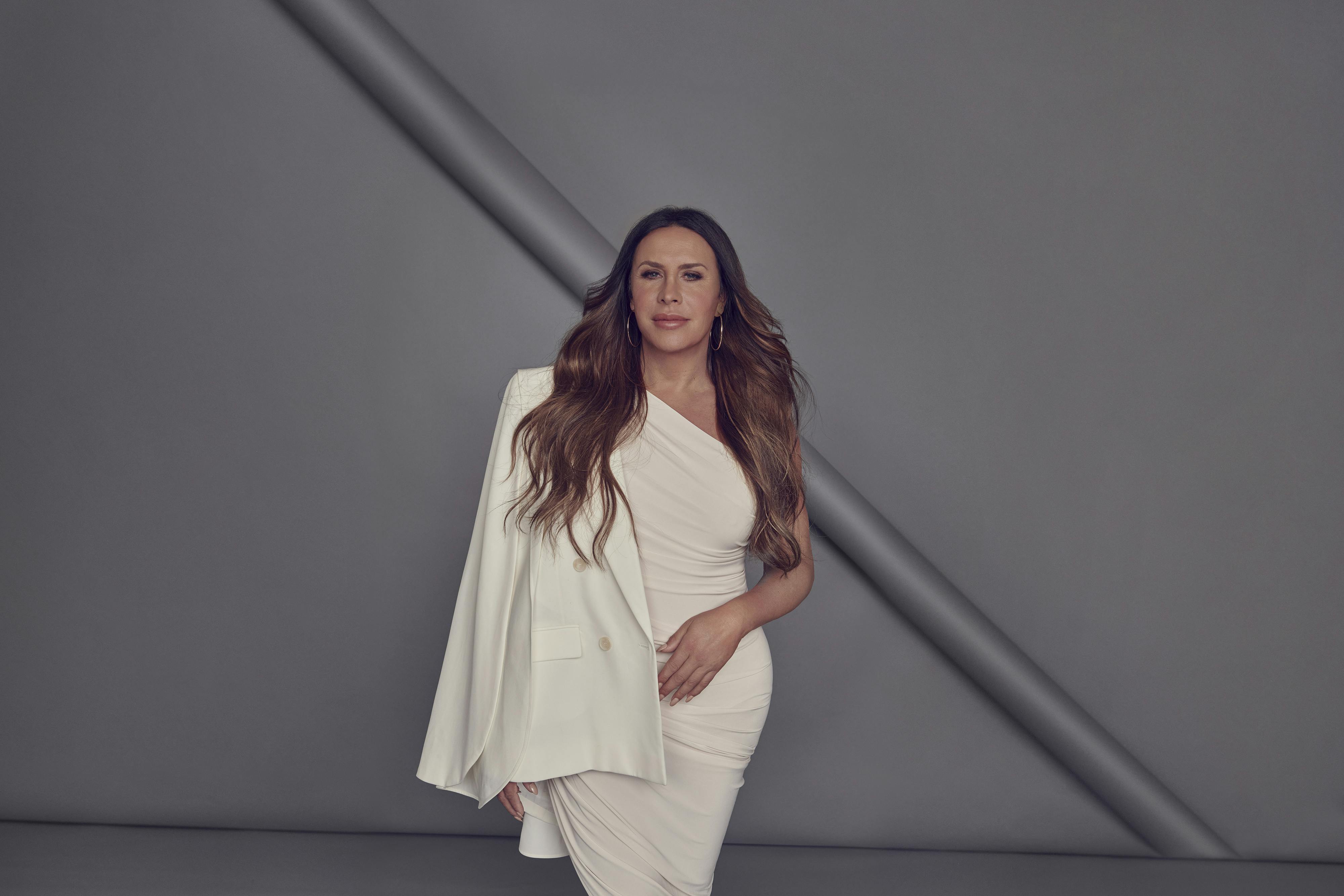 Karla Sofía Gascón wears a white one-shouldered dress against a grey background.