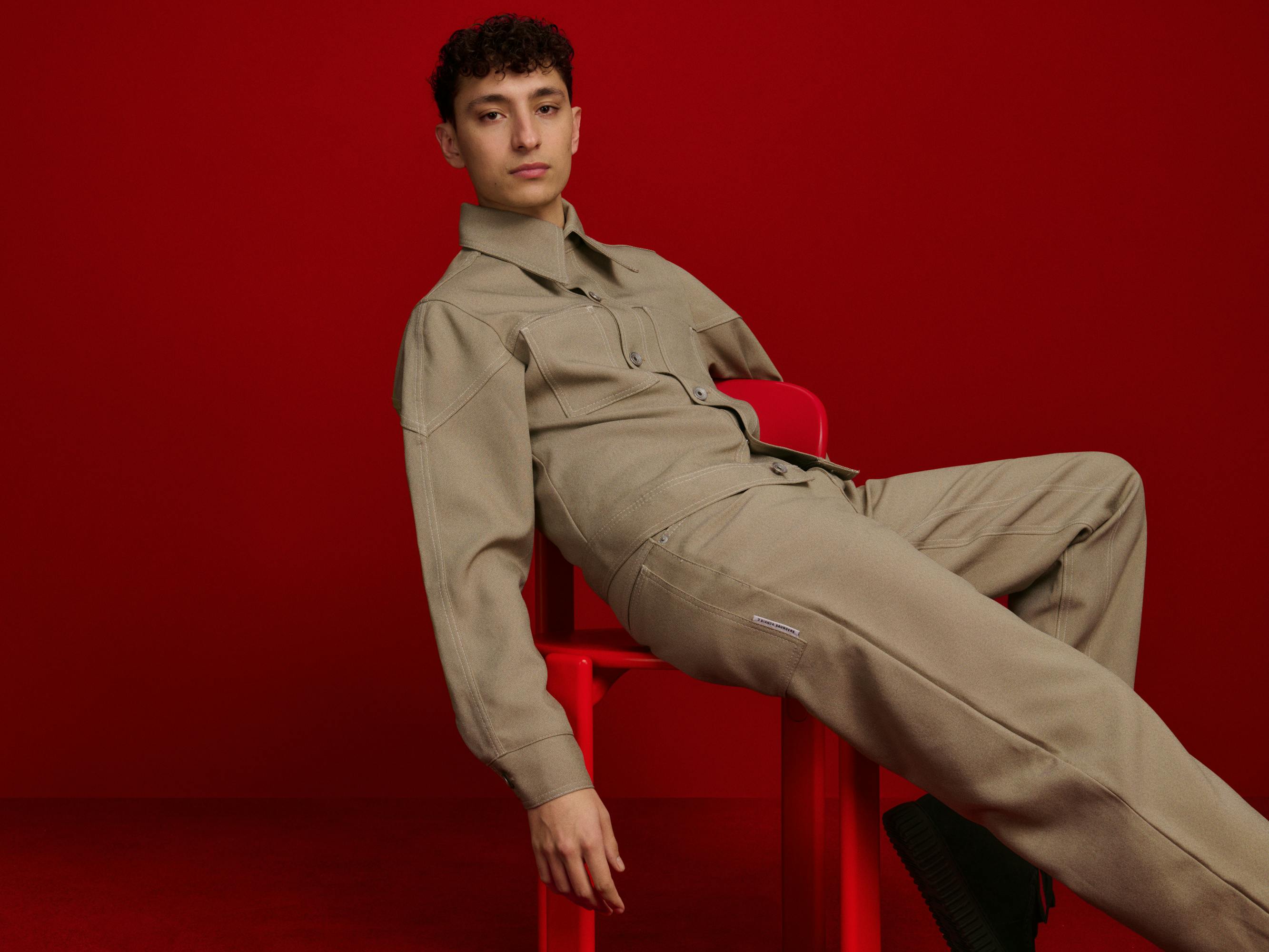 Jayden Revri wears a khaki suit against a a red background, sitting sideways in a red chair.