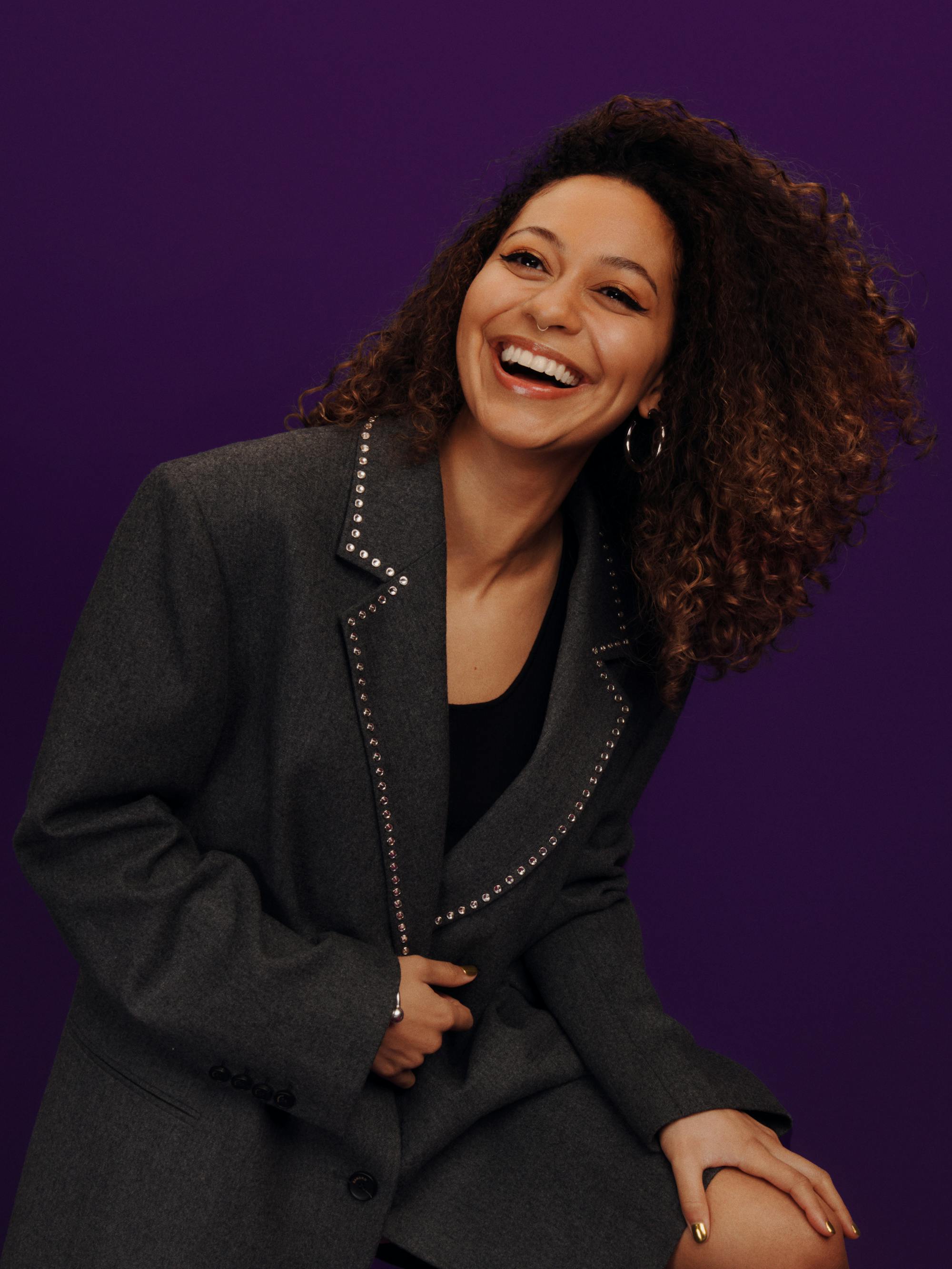 Kassius Nelson wears a black blazer and smiles wide in a purple room. 