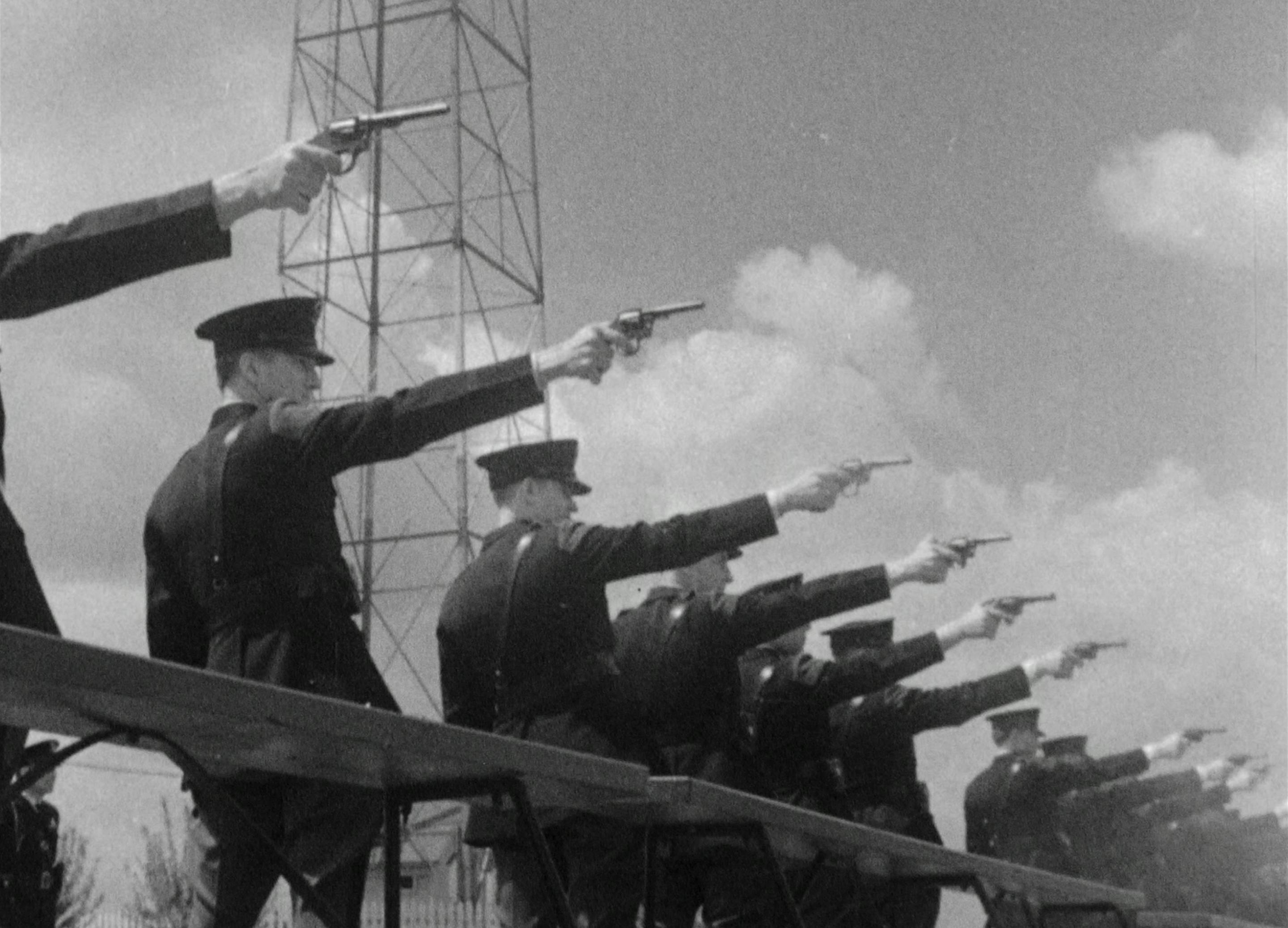 A still from the film: Police men standing in a row aim their guns at something in the distance.