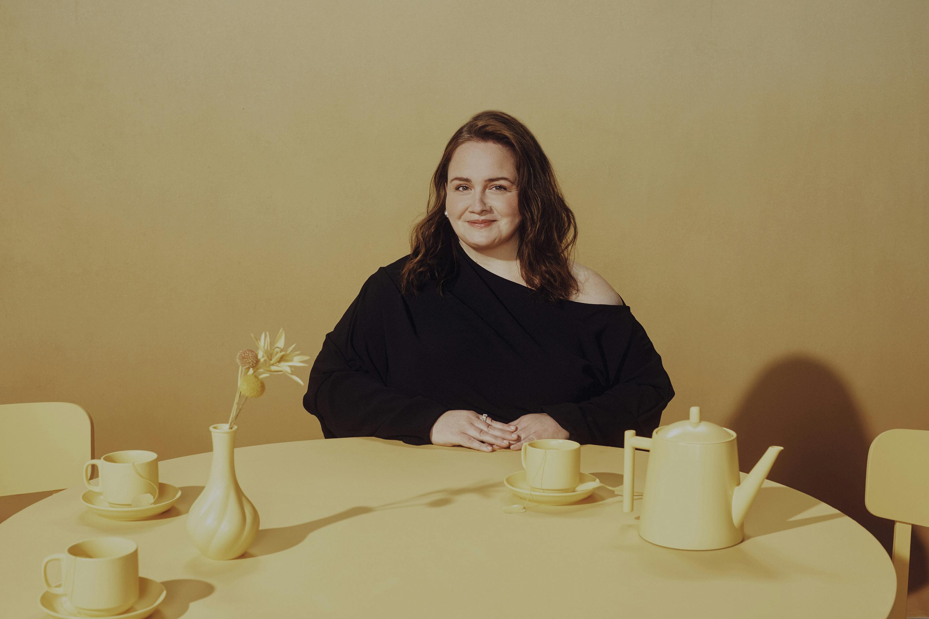 Jessica Gunning smiles knowingly in a black top, sitting at a yellow table with a yellow tea set and vase.