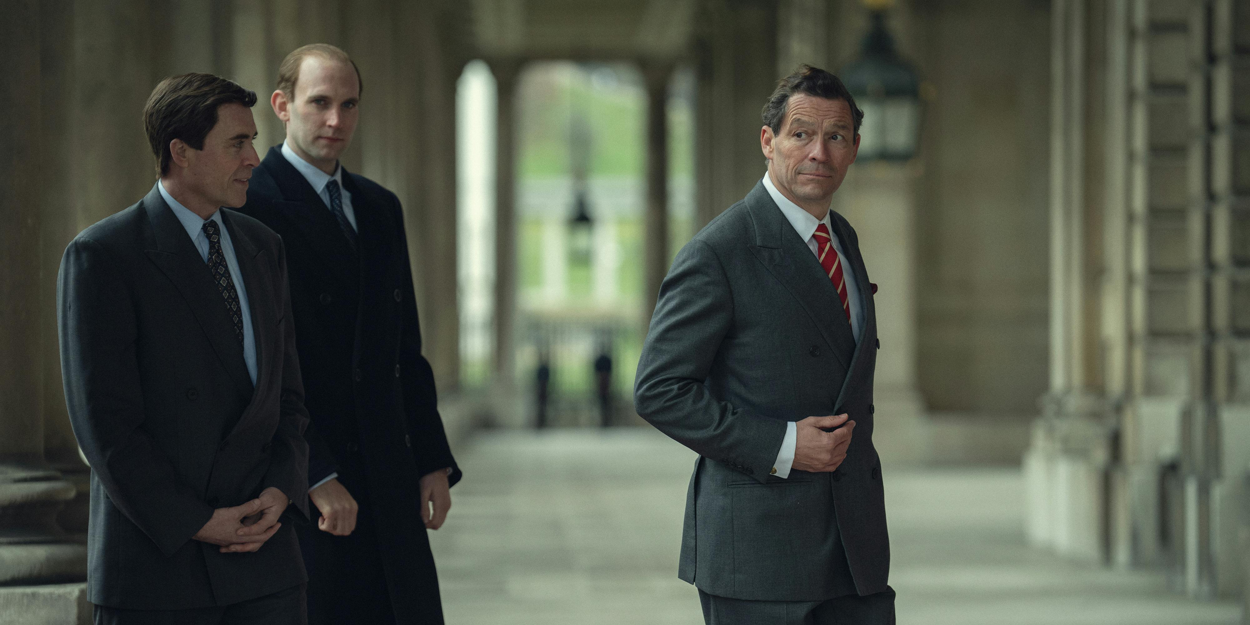 Prince Charles (Dominic West) wears a suit and red tie and walks through a airy hallway with some cronies.