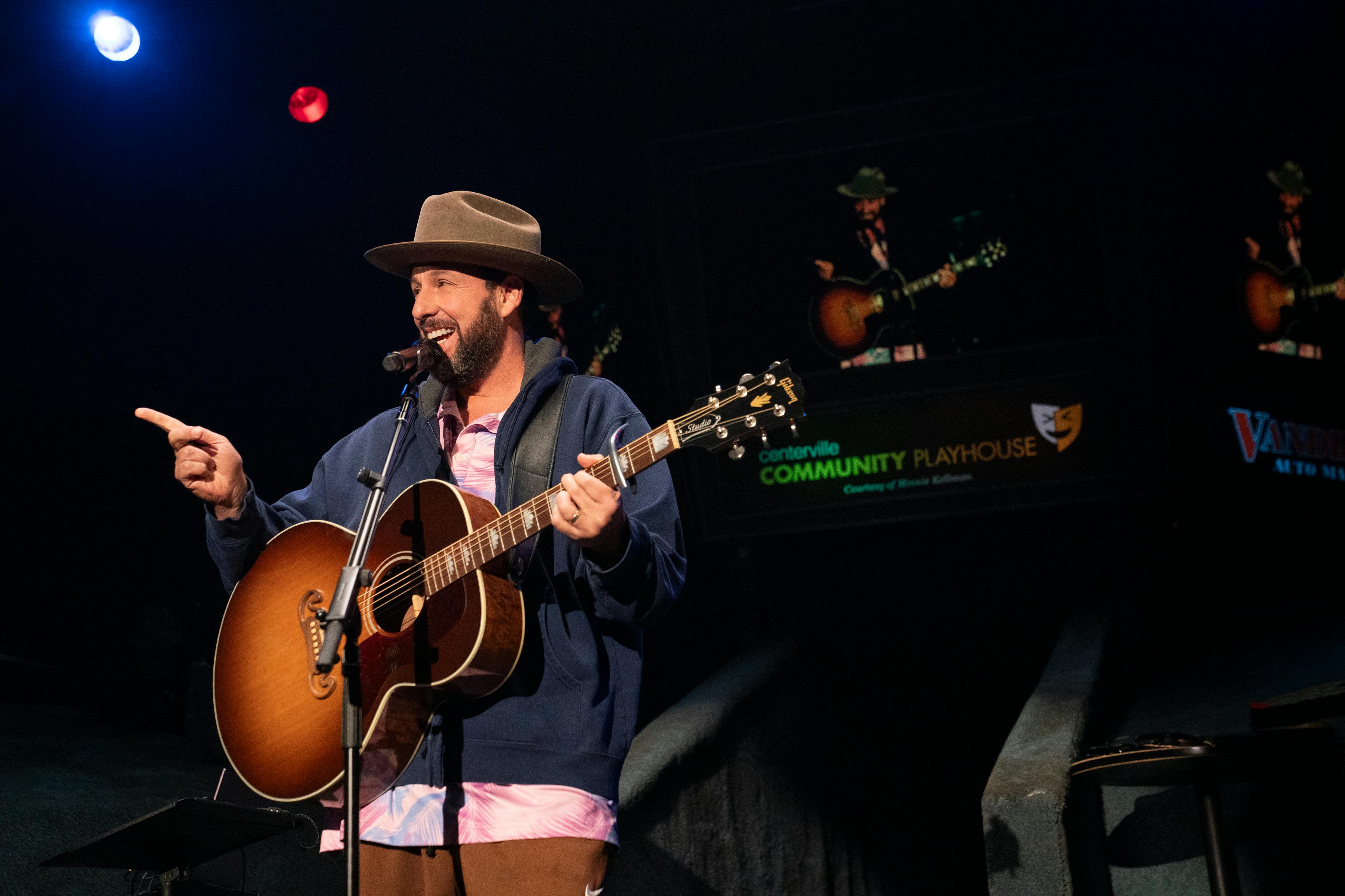 Adam Sandler wears a gray hat and plays guitar on a darkened stage.