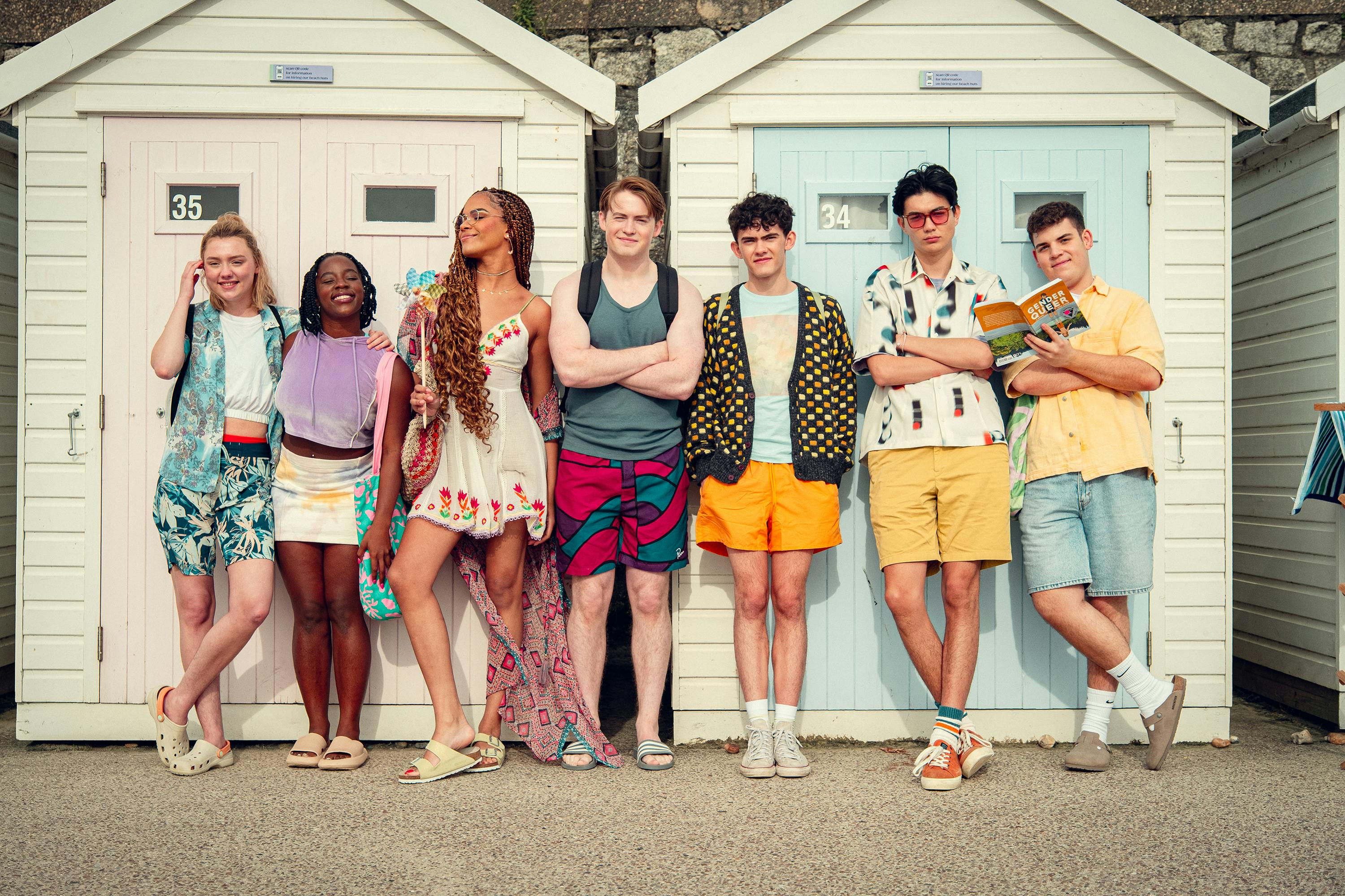 The cast of Heartstopper line up against some white cottages looking beachy and adorable.