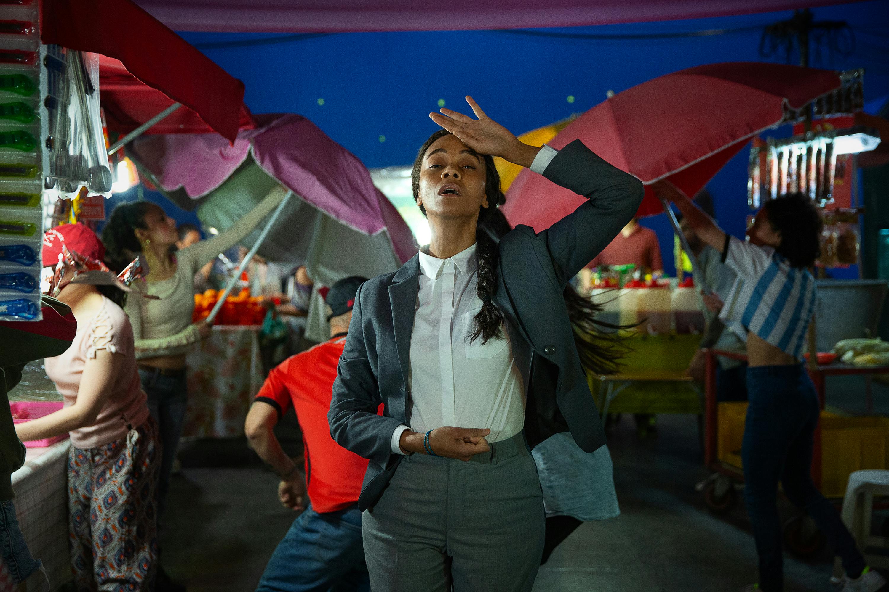 Rita (Zoe Saldaña) brings the back of her hand to her forehead in this dramatic shot in an outdoor street market.