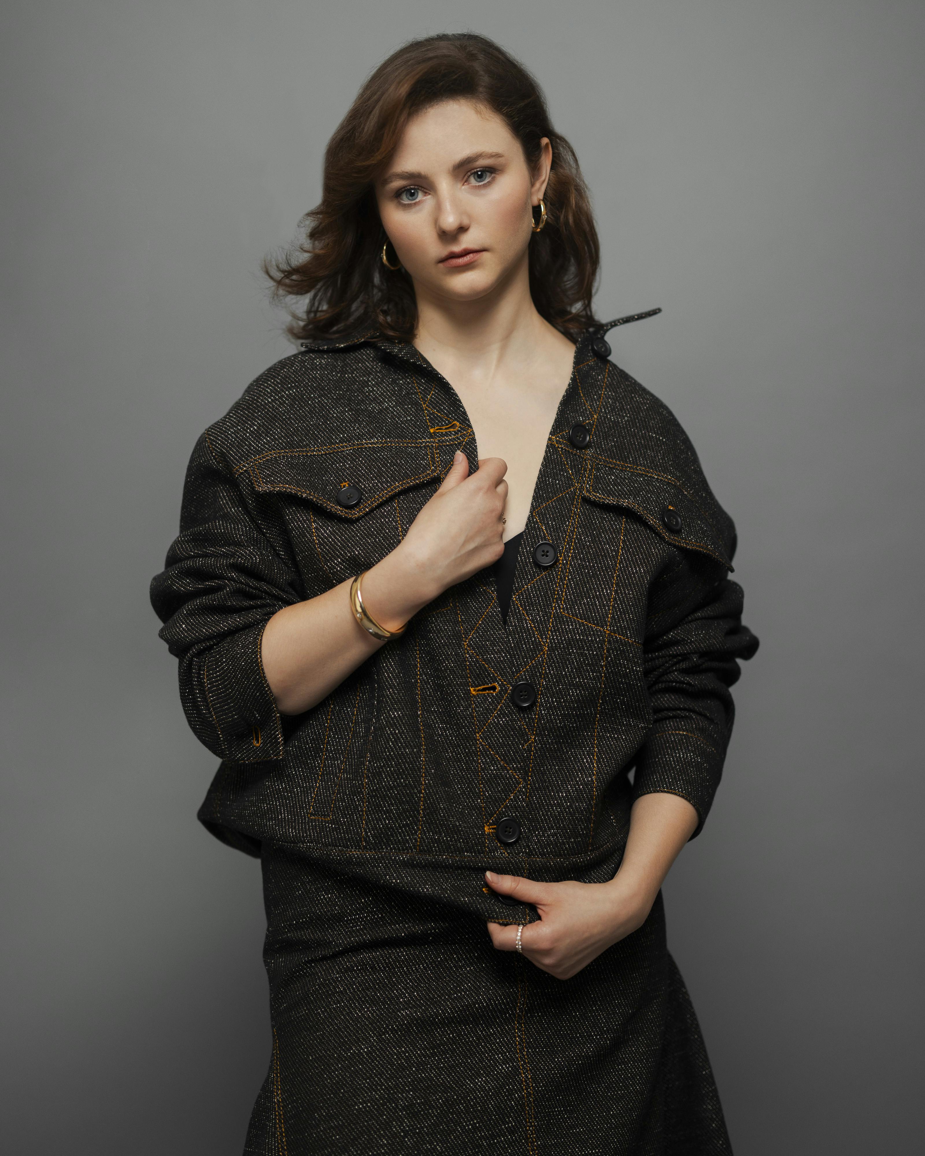 Thomasin McKenzie stands in front of a grey background looking to camera in a matching denim jacket and skirt set.