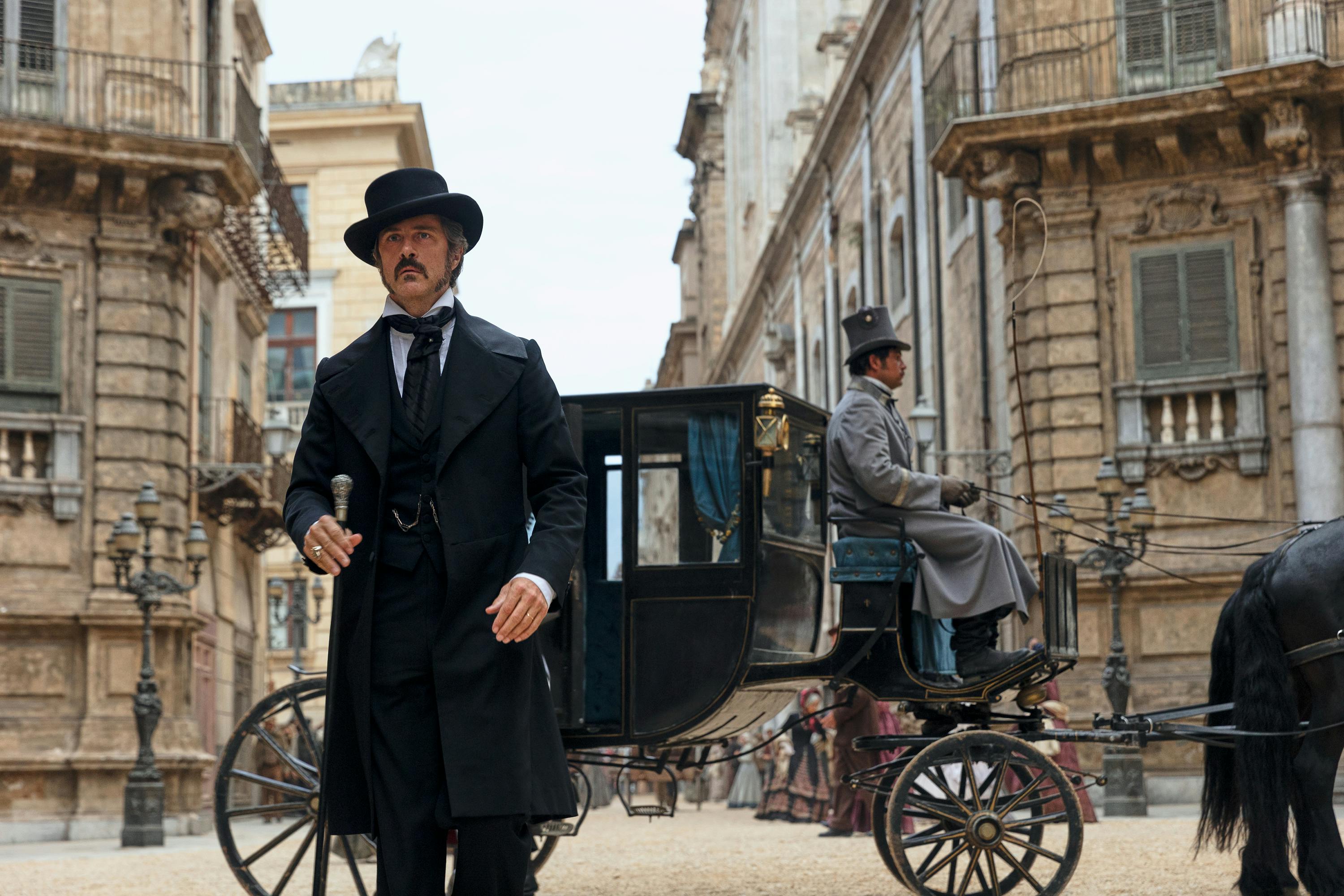 A scene from The Leopard. A man wears a morning coat and top hat and stands outside a horse and buggy. 