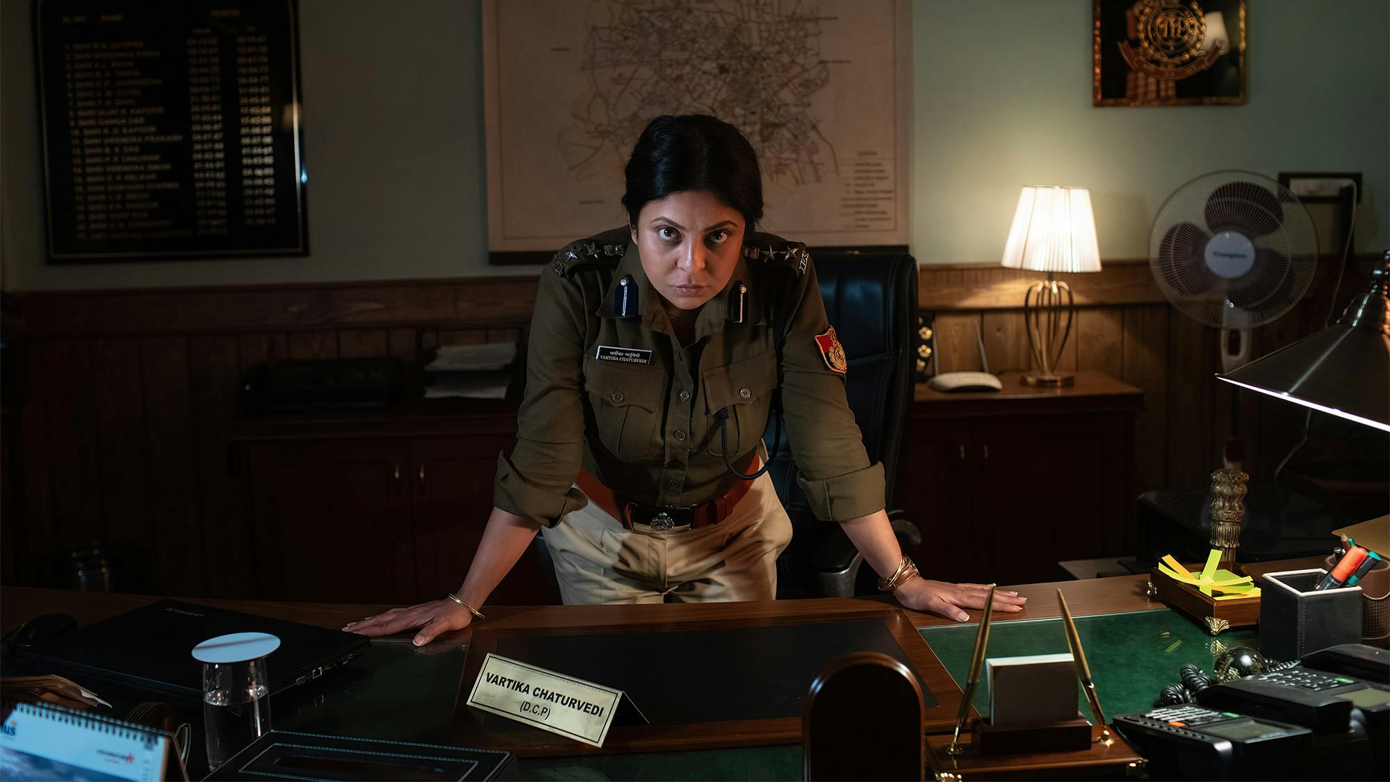 Shefali Shah as Vartika Chaturvedi stands in her police uniform, leaning over onto her desk. One light is on behind her.  