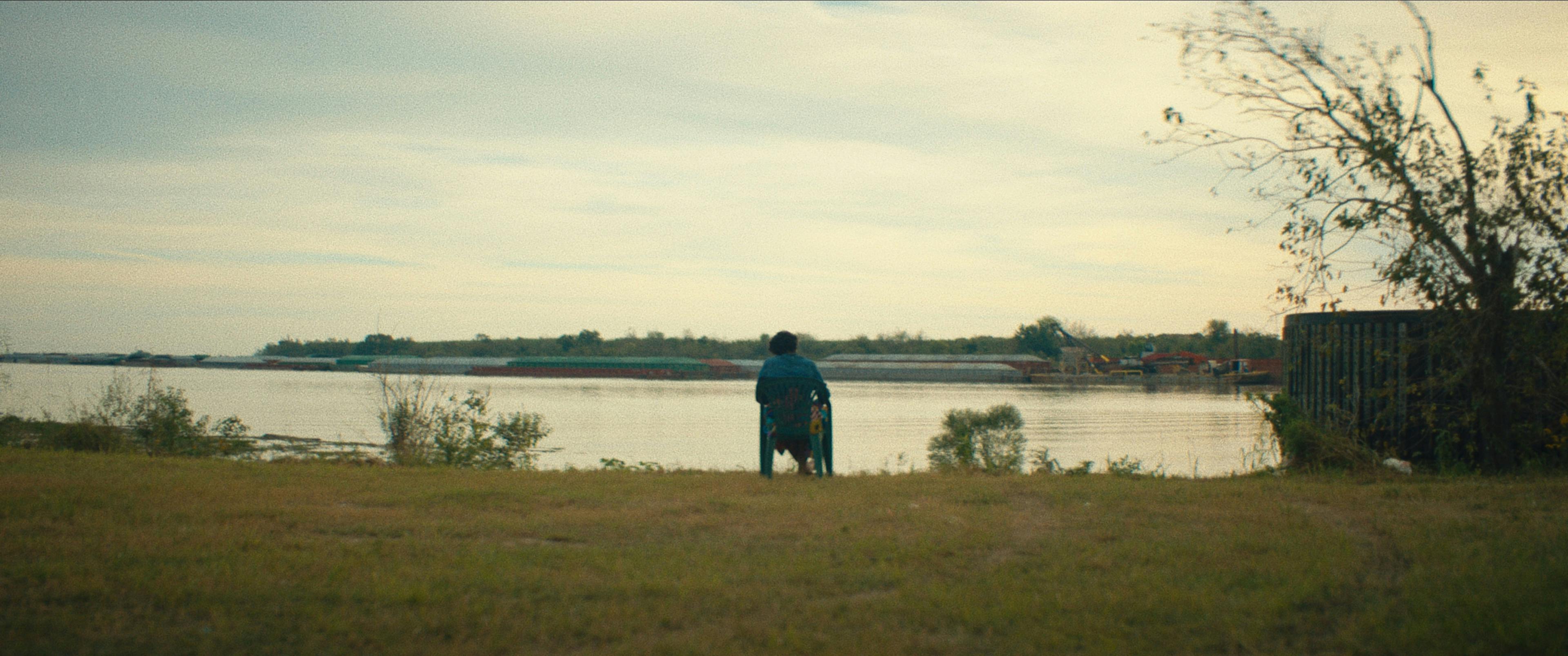 A still from descendant. Someone sits on a chair looking out onto a river.