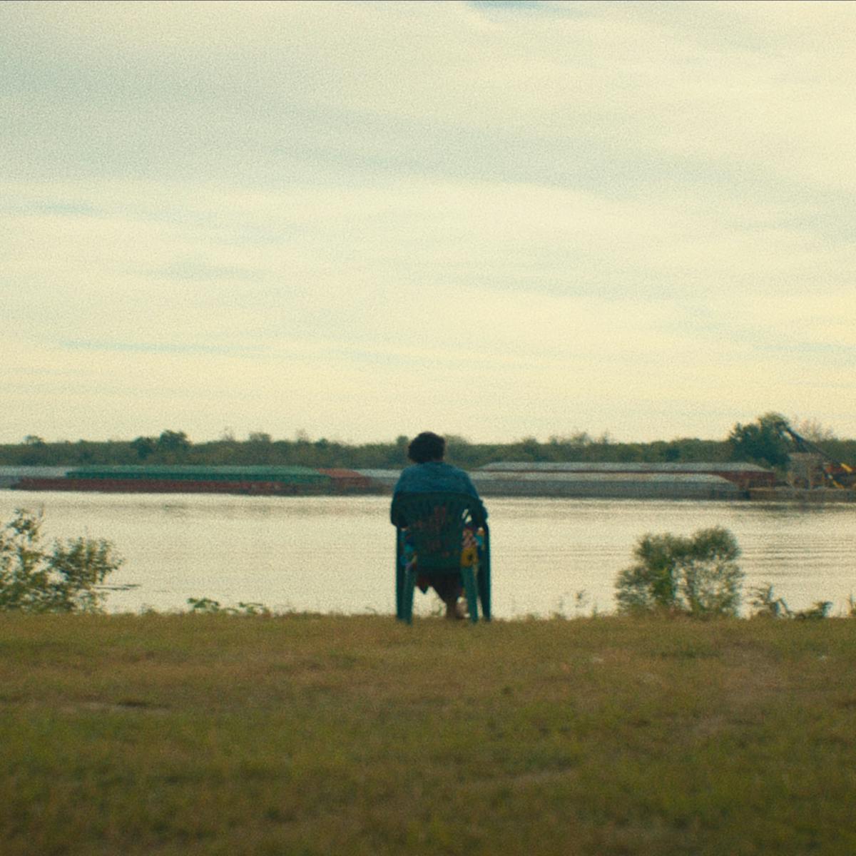 A still from descendant. Someone sits on a chair looking out onto a river.