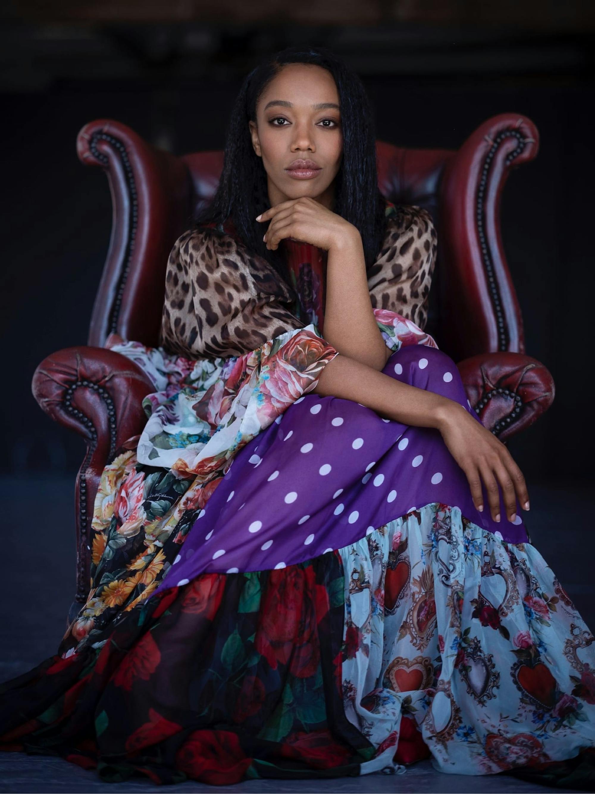 Noami Ackie wears the same patchwork dress as the opening shot, but this time she sits in a red leather chair. She rests her chin on her hand, and looks directly at the camera. The background is dark and shadowy.