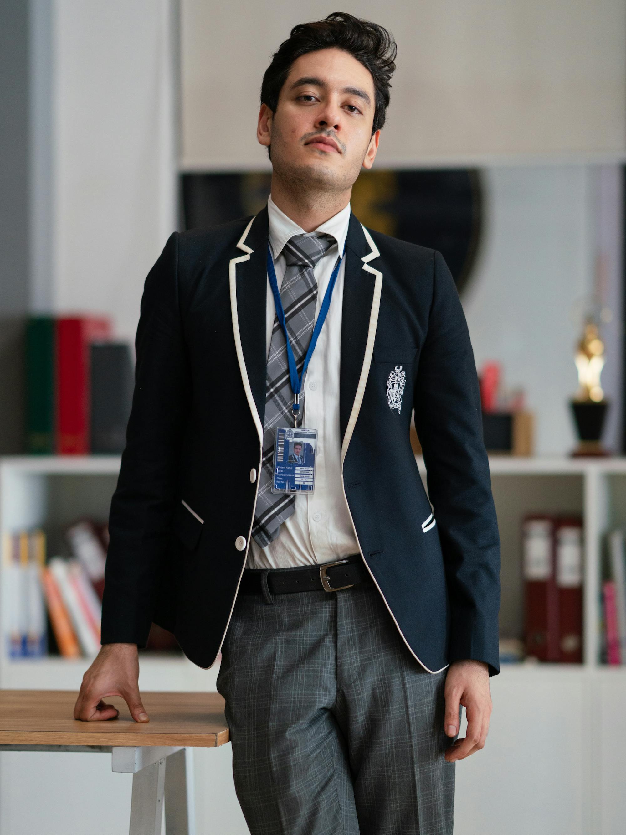 Veer (Zeyn Shaw) wears his school uniform: grey pants, a navy blazer, white shirt, grey plaid tie, and blue lanyard. 
