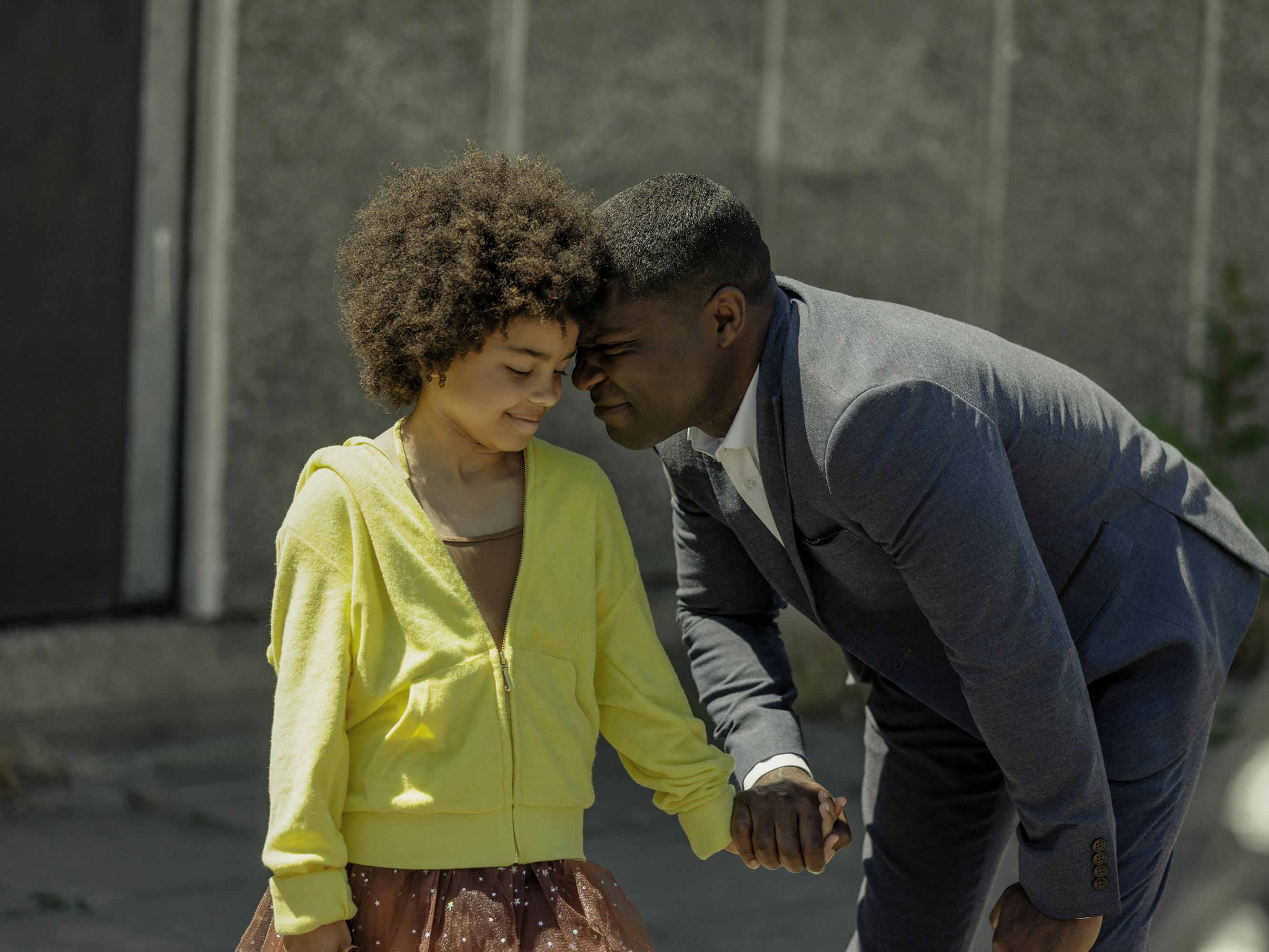 Laura (Amelie Dokubo) wears a neon yellow hoodie over a brown dress. Dayo (David Oyelowo) leans over and holds Laura’s hand.