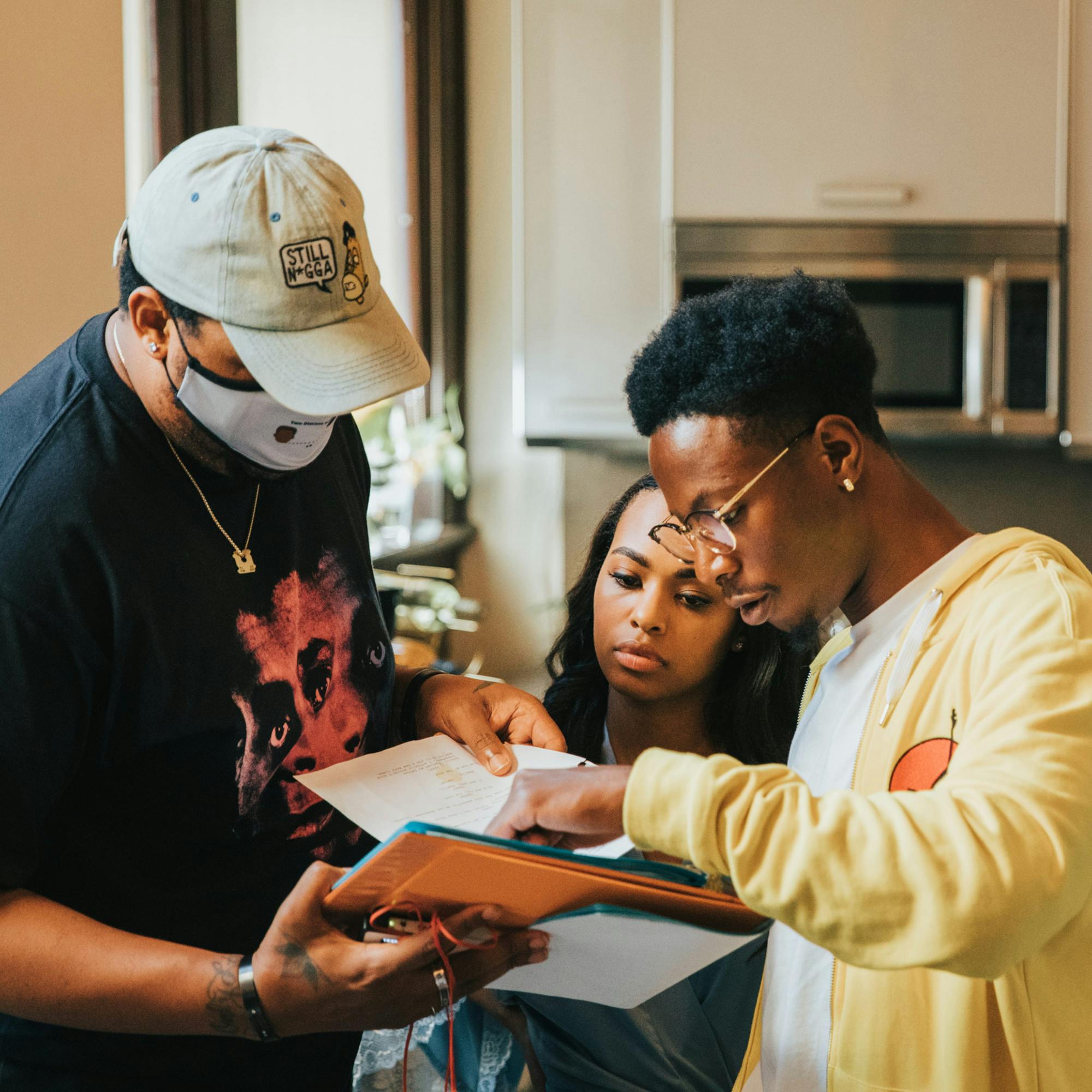 Free, Zaria, and Bada$$ review something in a folder. Zaria and Bada$$ look very focused, and Bada$$ wears his character’s trademarked yellow hoodie and glasses. Free wears a tan hat, white mask, and a black shirt with faces on it. 