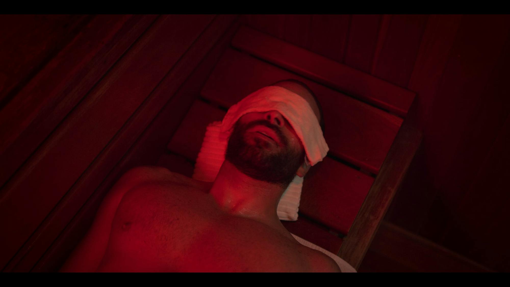 Rodolfo (Alejandro Nones) lies on a wooden bench in the sauna with a towel over his eyes and behind his head. The red lighting sets an ominous glow.