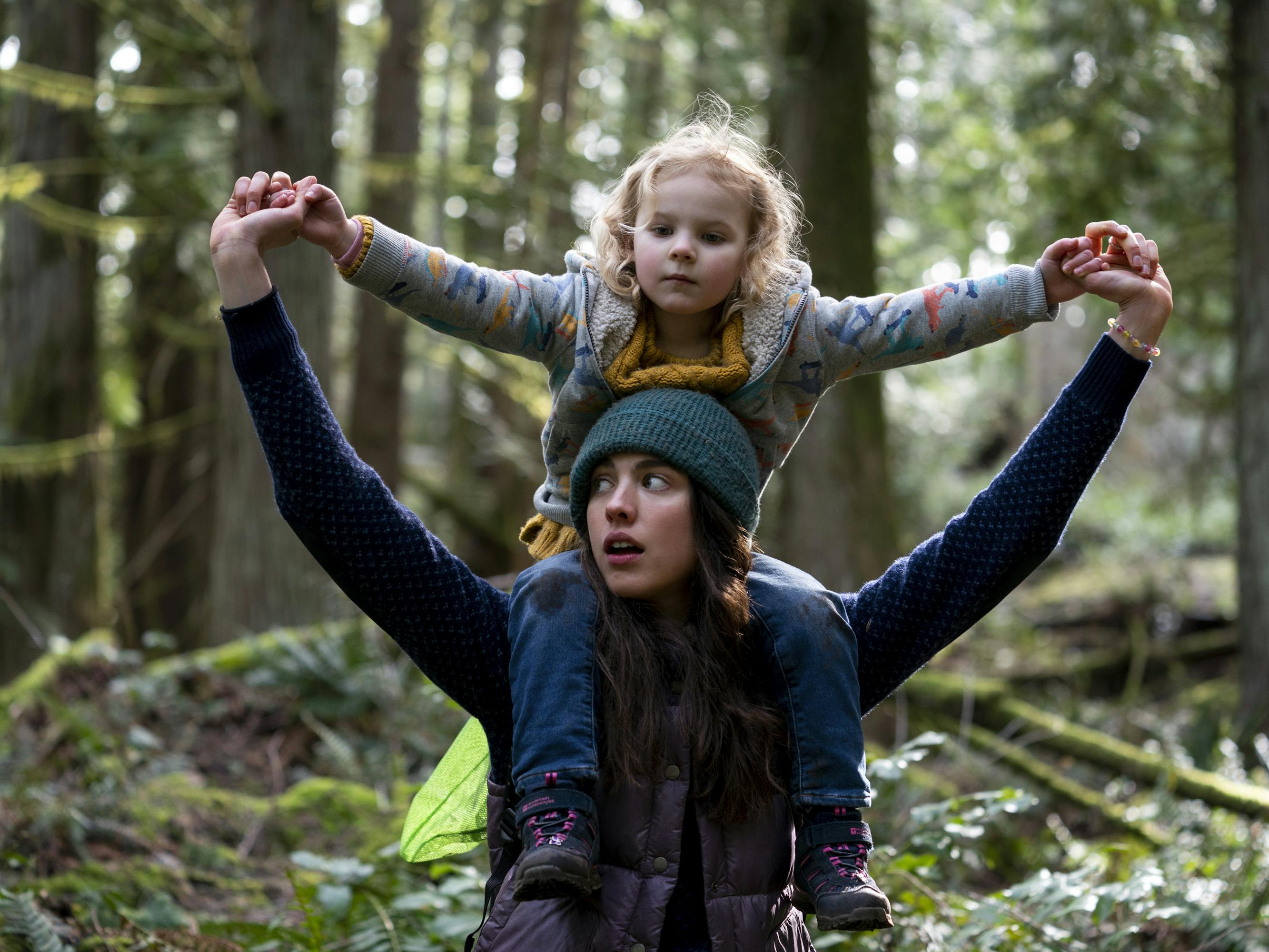 Maddy (Rylea Nevaeh Whittet) and Alex Russell (Margaret Qualley) are going on a bear hunt! Maddy sits on her moms shoulders, and sunlight peeks through the green trees.