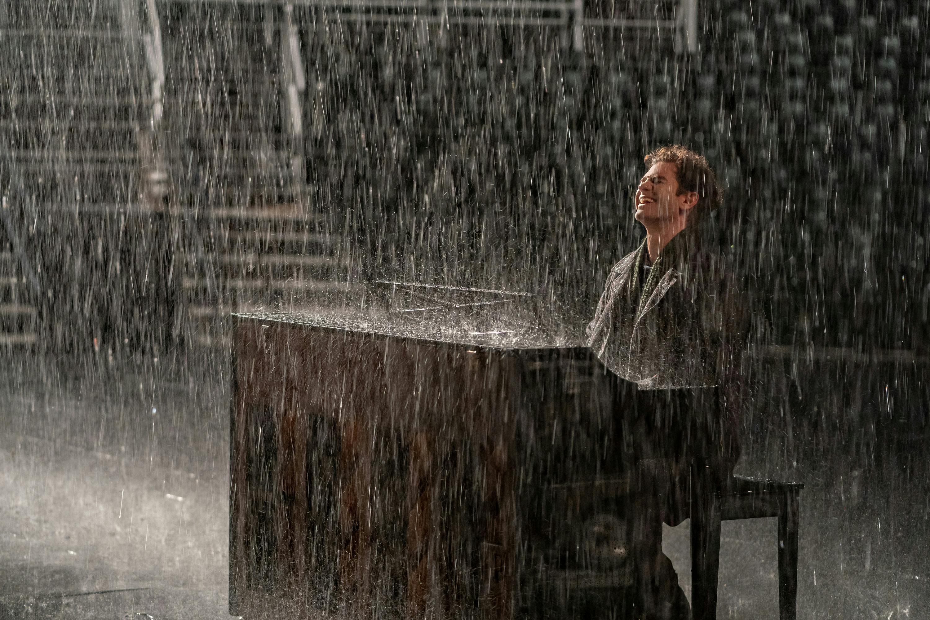 Andrew Garfield plays piano outside in a massive rainstorm. He wears dark clothing and smiles wide.