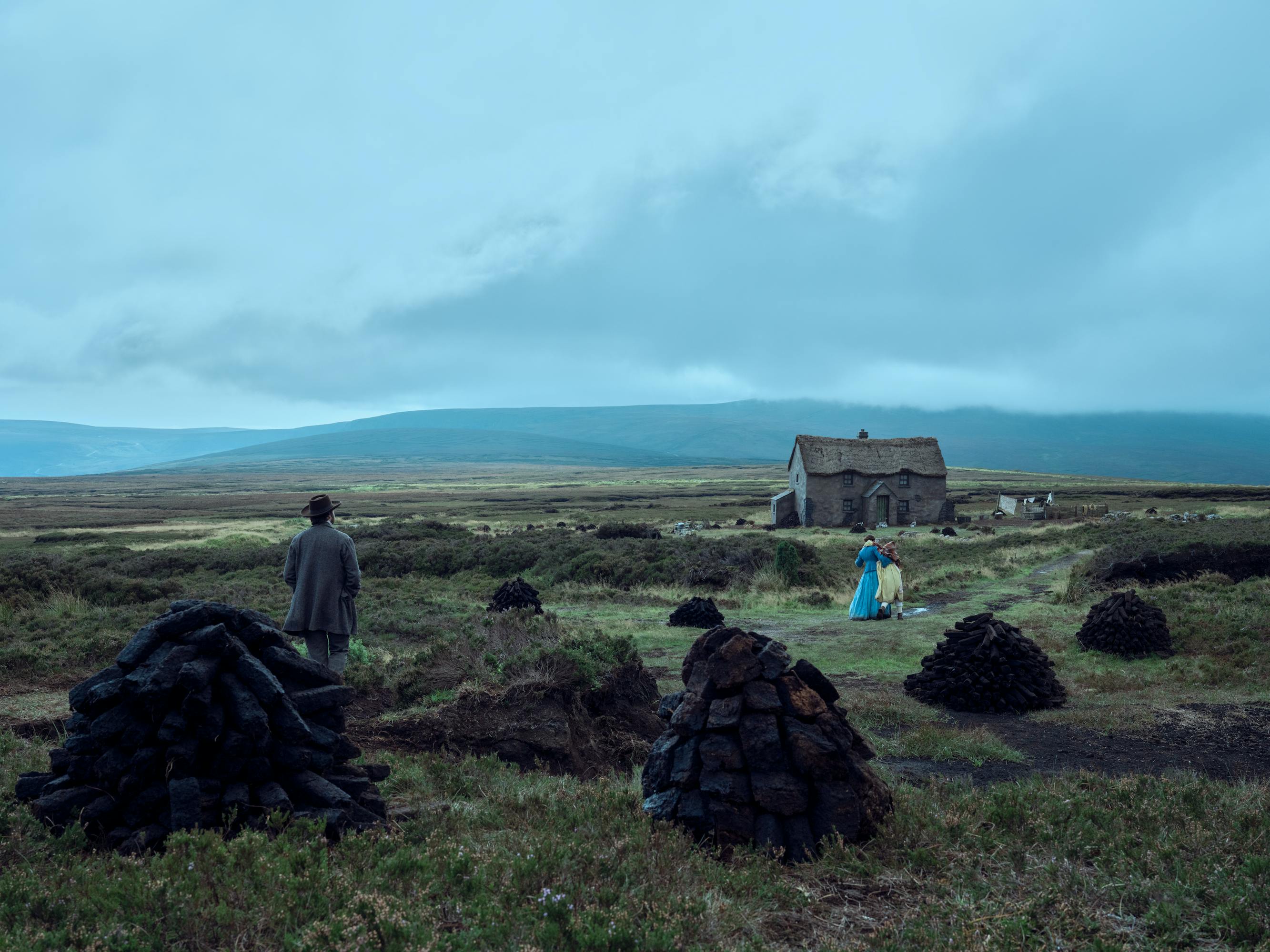 Tom Burke, Florence Pugh, and Kíla Lord Cassidy make there way to a sunken looking house in a grey landscape.