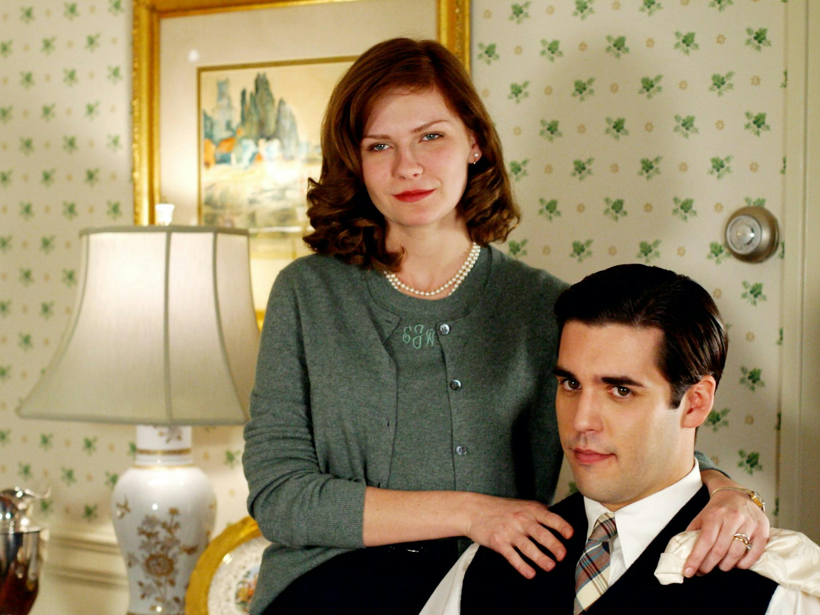 Betty Warren (Kirsten Dunst) stands behind a sitting Spencer Jones (Jordan Bridges) in a 1950s style home. Dunst wears a green sweater set and Bridges wears a black vest, white shirt, and tie.