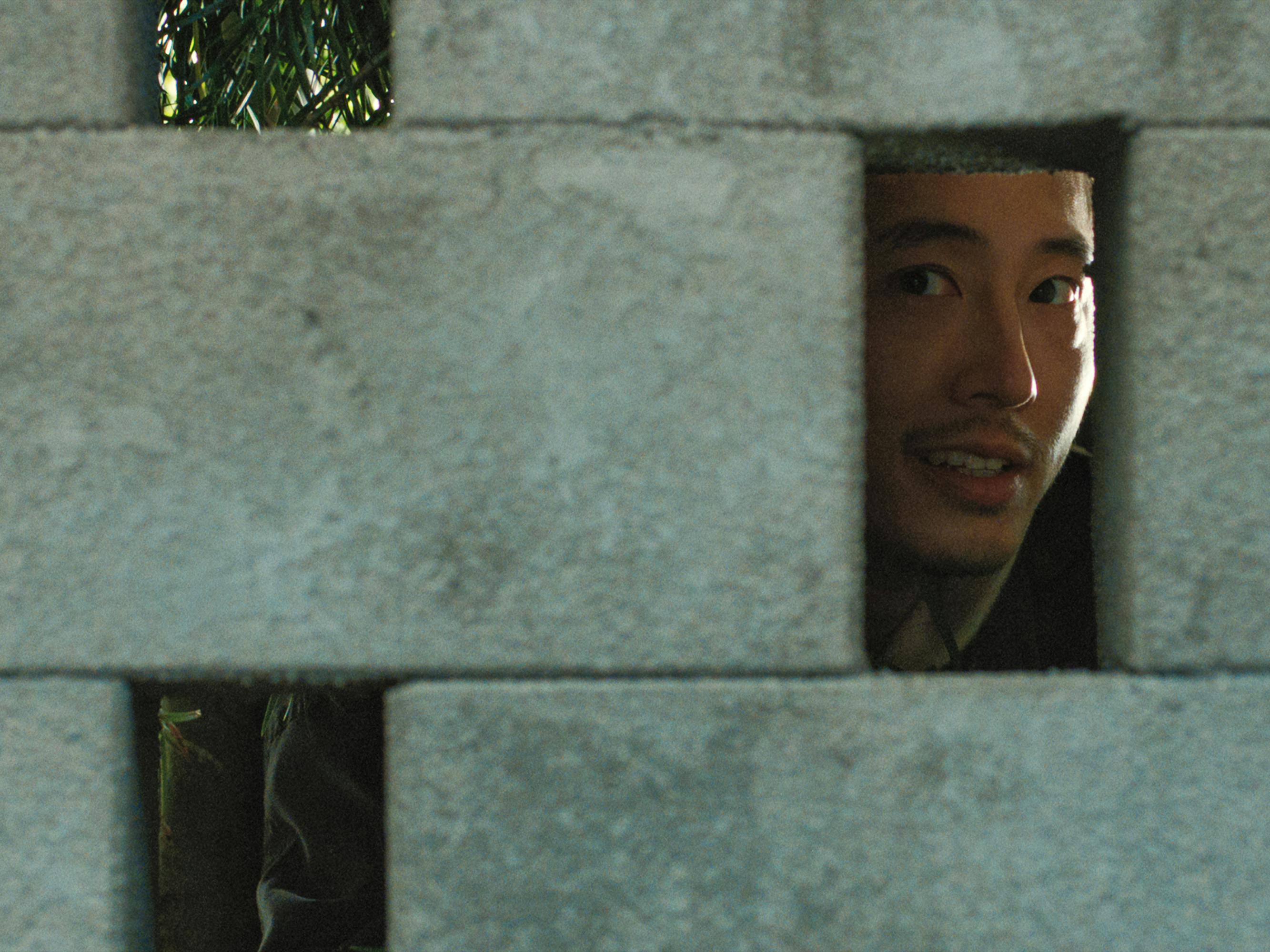 Steven Yeun looks through a crack in a stone wall.