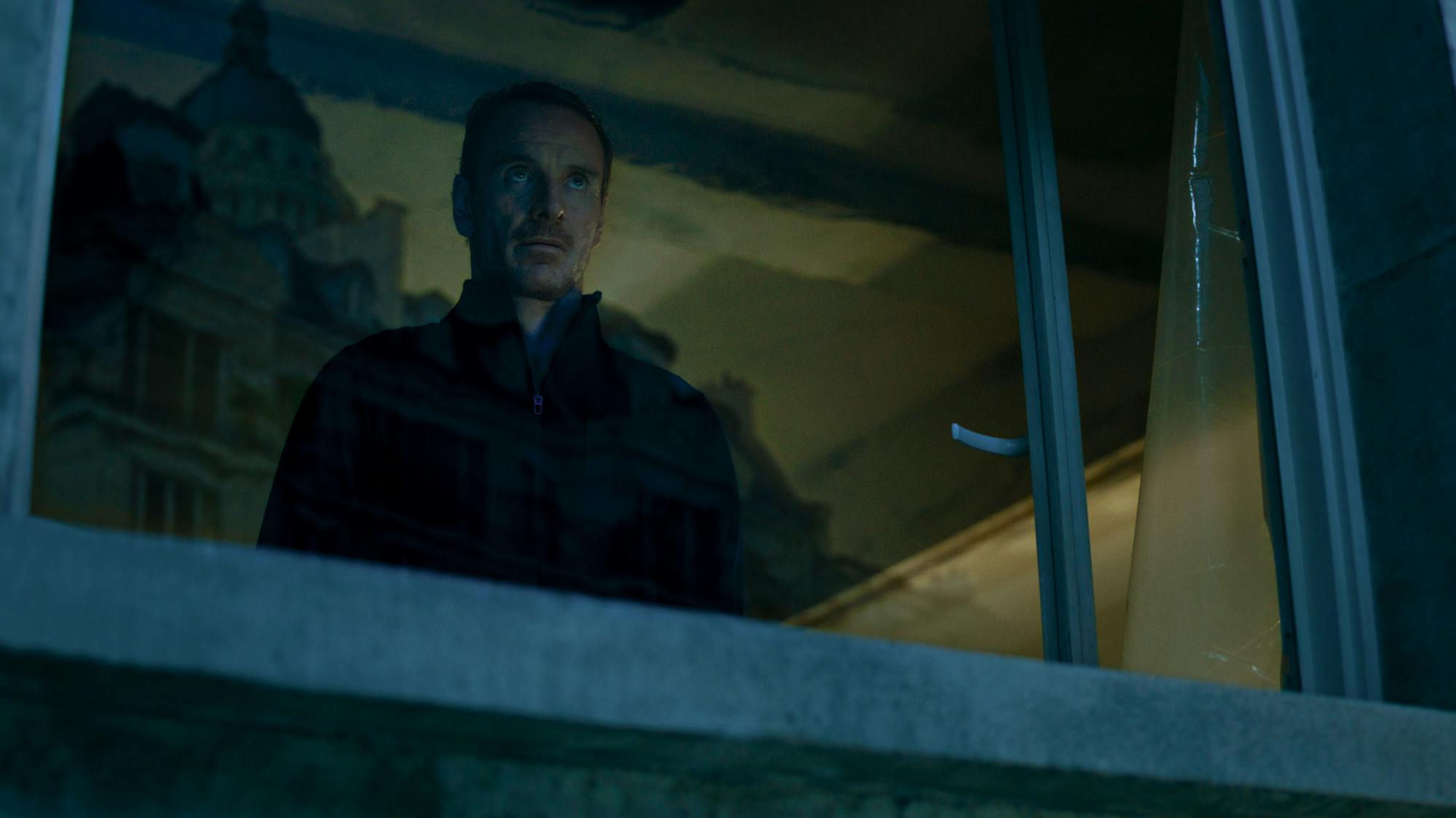 The Killer (Michael Fassbender) looks through a window. He wears all black and looks intense!