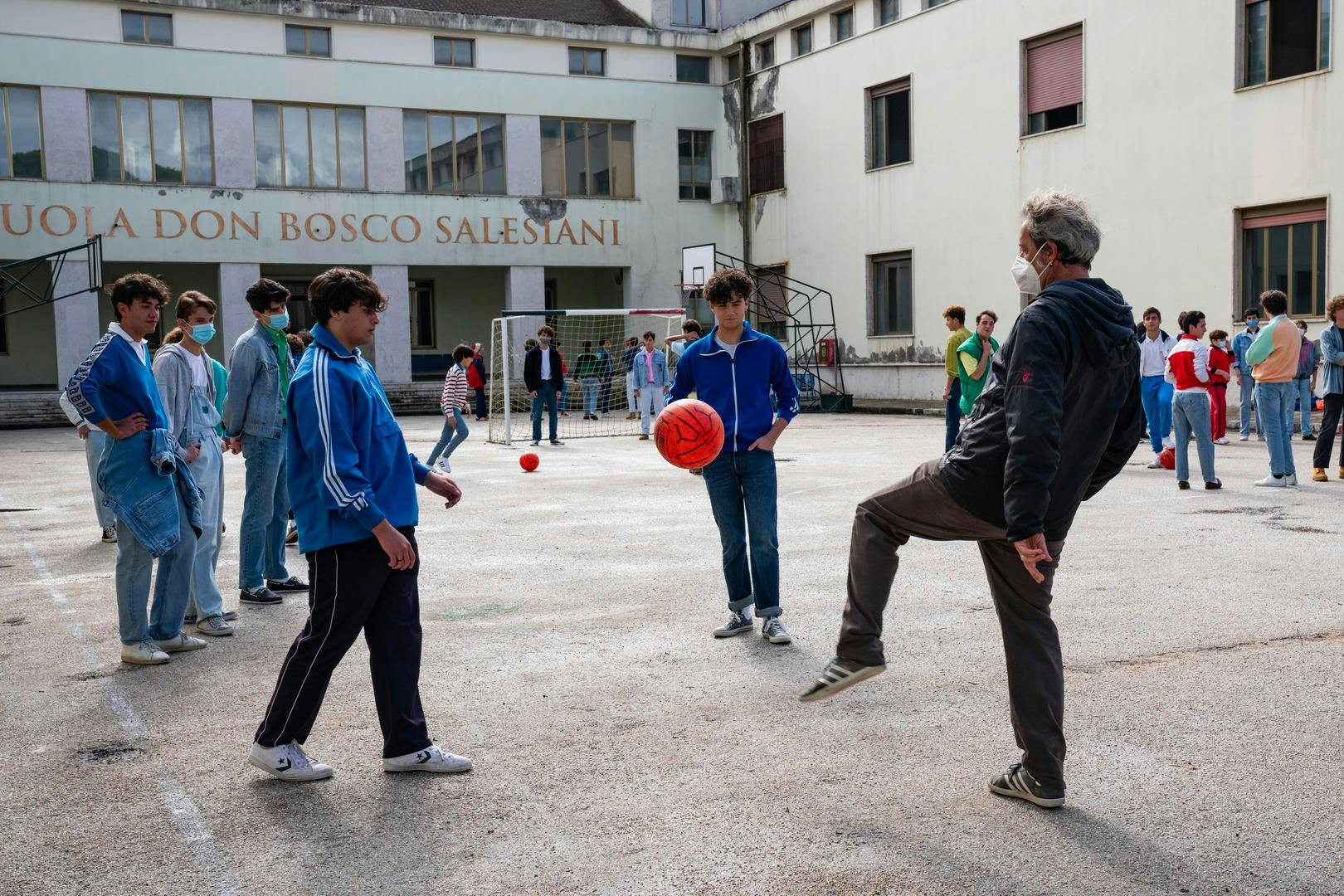 A scene of boys and Paolo Sorrentino behind a cement building. Many of the boys wear blue track jackets and most of the figures are masked. Sorrentino kicks an orange ball towards one of the other boys.