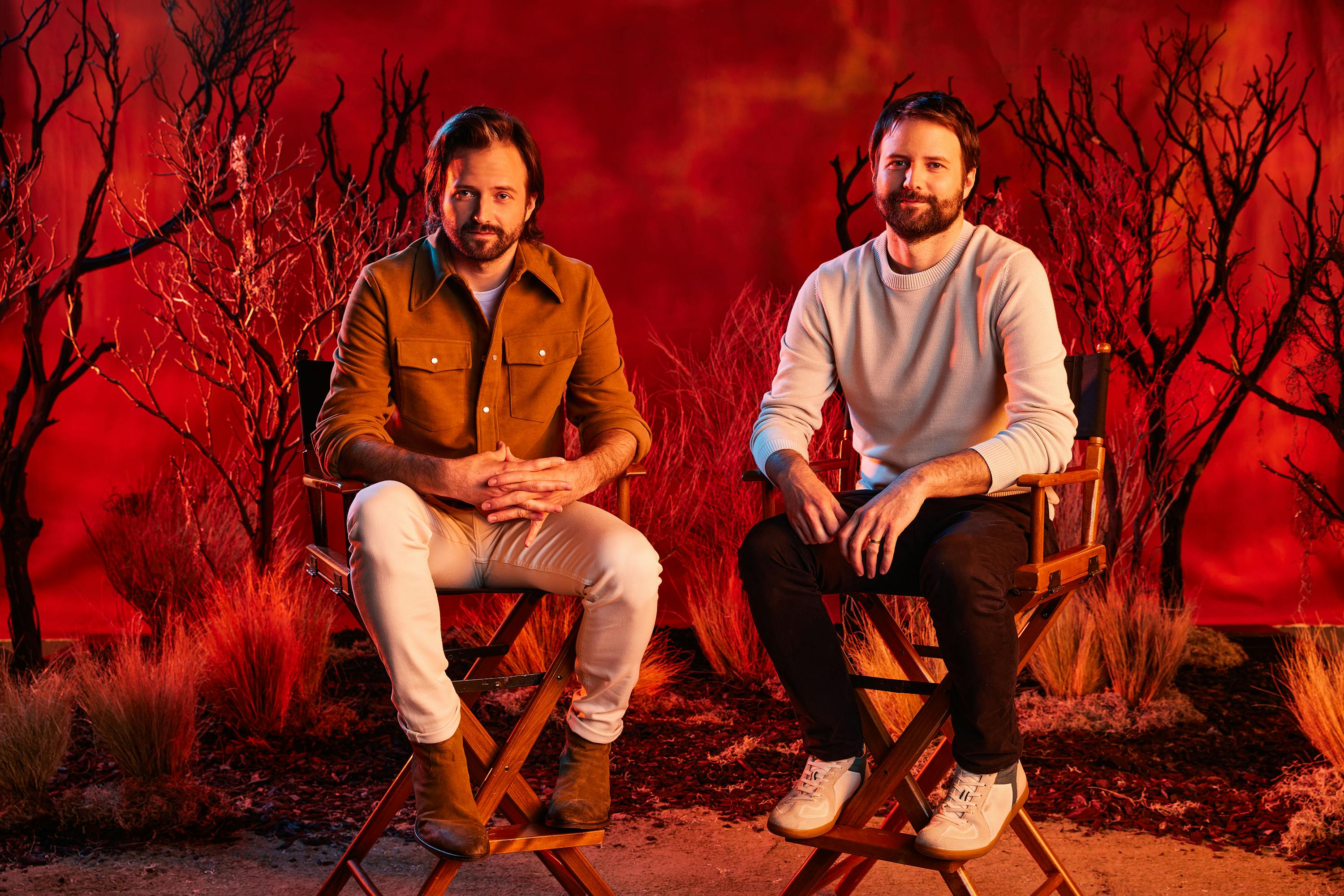 Matt and Ross Duffer sit in directors chairs against a fiery red backdrop.