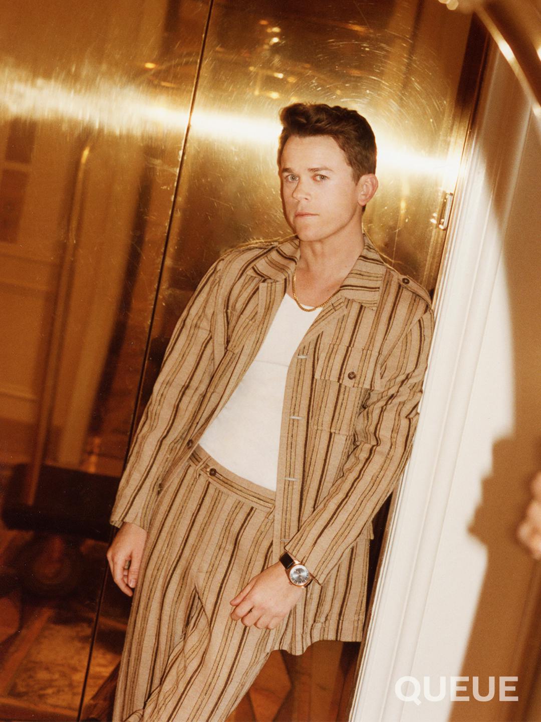 John Owen Lowe wears a striped brown suit over a white tank top and leans against white molding.