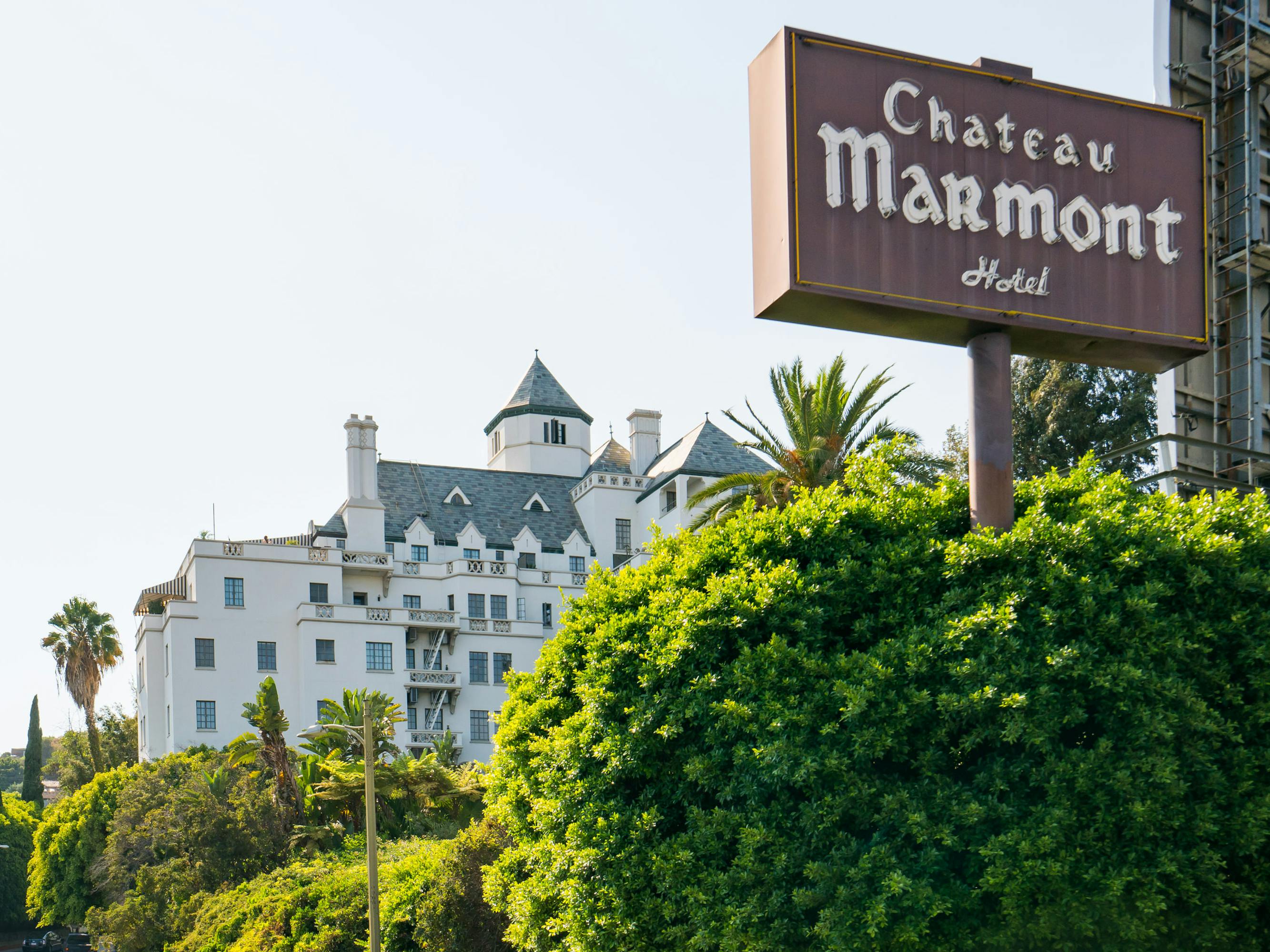 Chateau Marmont, as announced by a brown sign with white lettering. The hotel is castle like, white with gray roofing. 