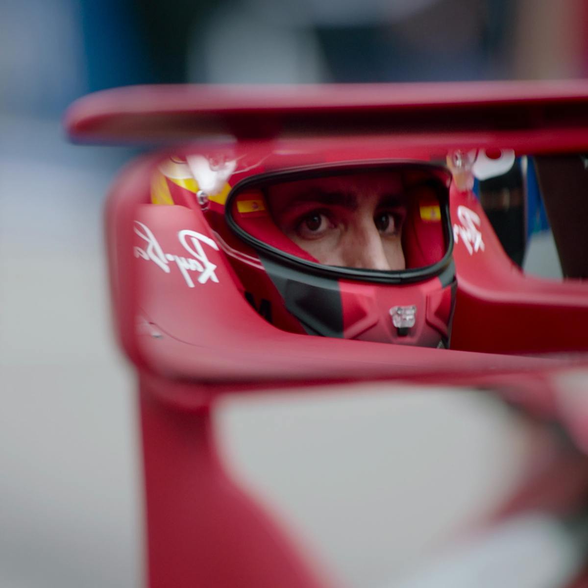 Driver Carlos Sainz Jr. sits in his red Ferrari F1 car looking into his rearview mirror.