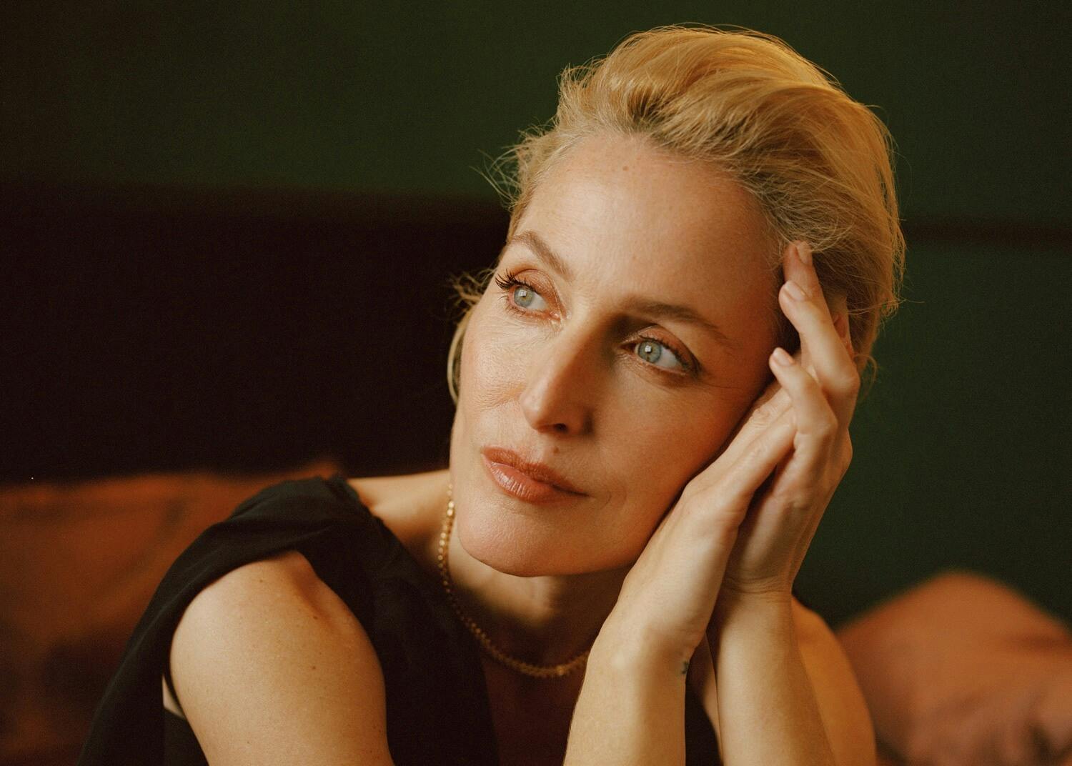 Gillian Anderson rests her head against her hands. She wears a black top and gold necklace and looks at something in the distance.