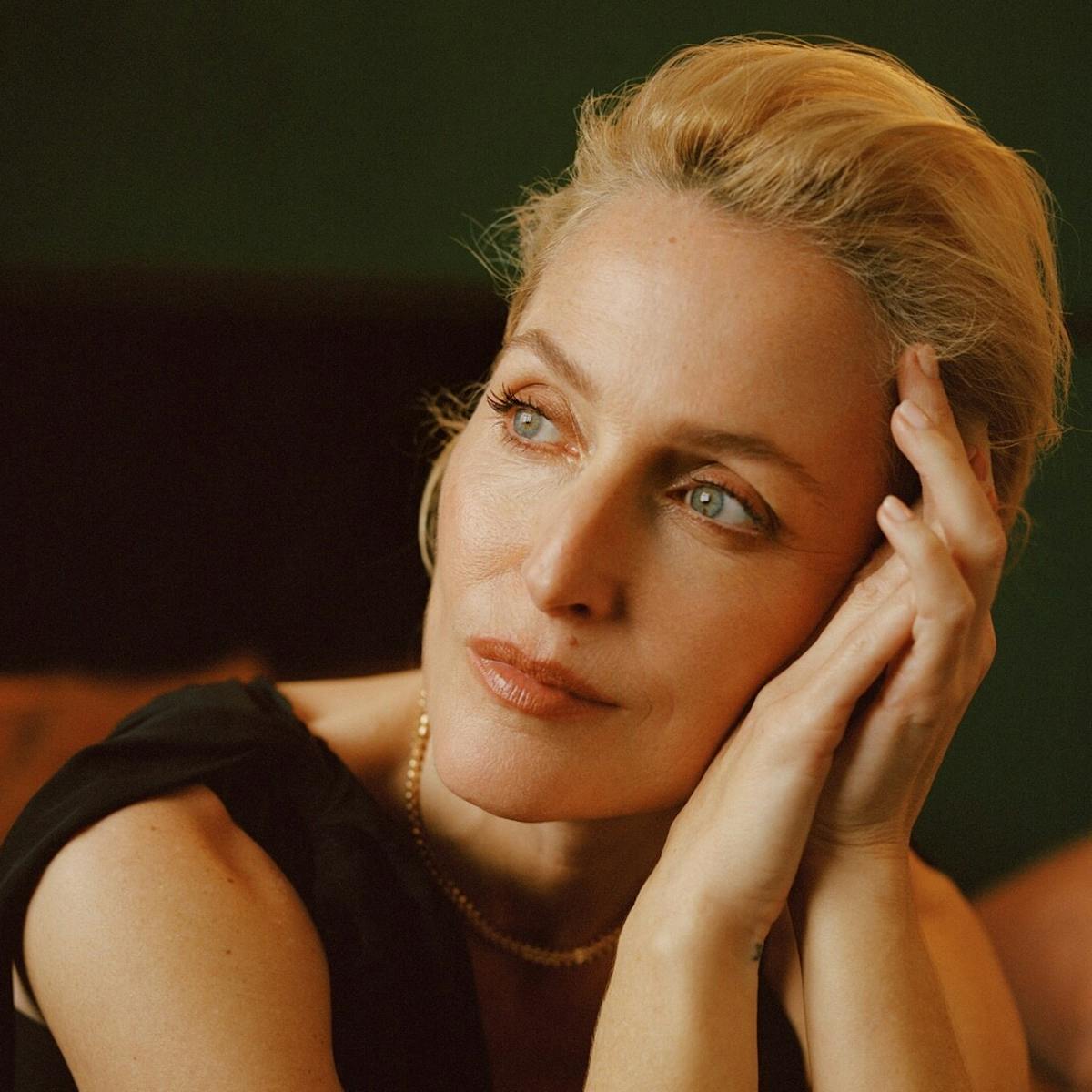 Gillian Anderson rests her head against her hands. She wears a black top and gold necklace and looks at something in the distance.