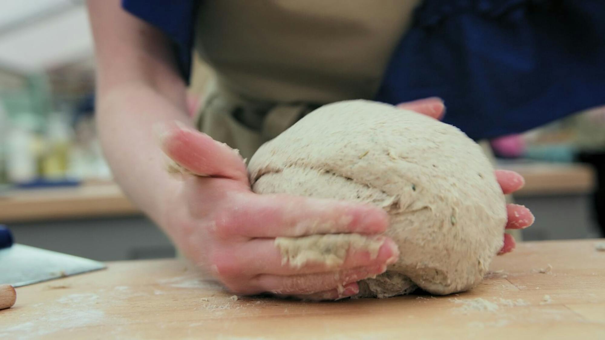 A pair of hands knead a ball of dough. The shot gives the sense of movement, motion, and speed. The person to whom the hands belong wears a khaki apron.