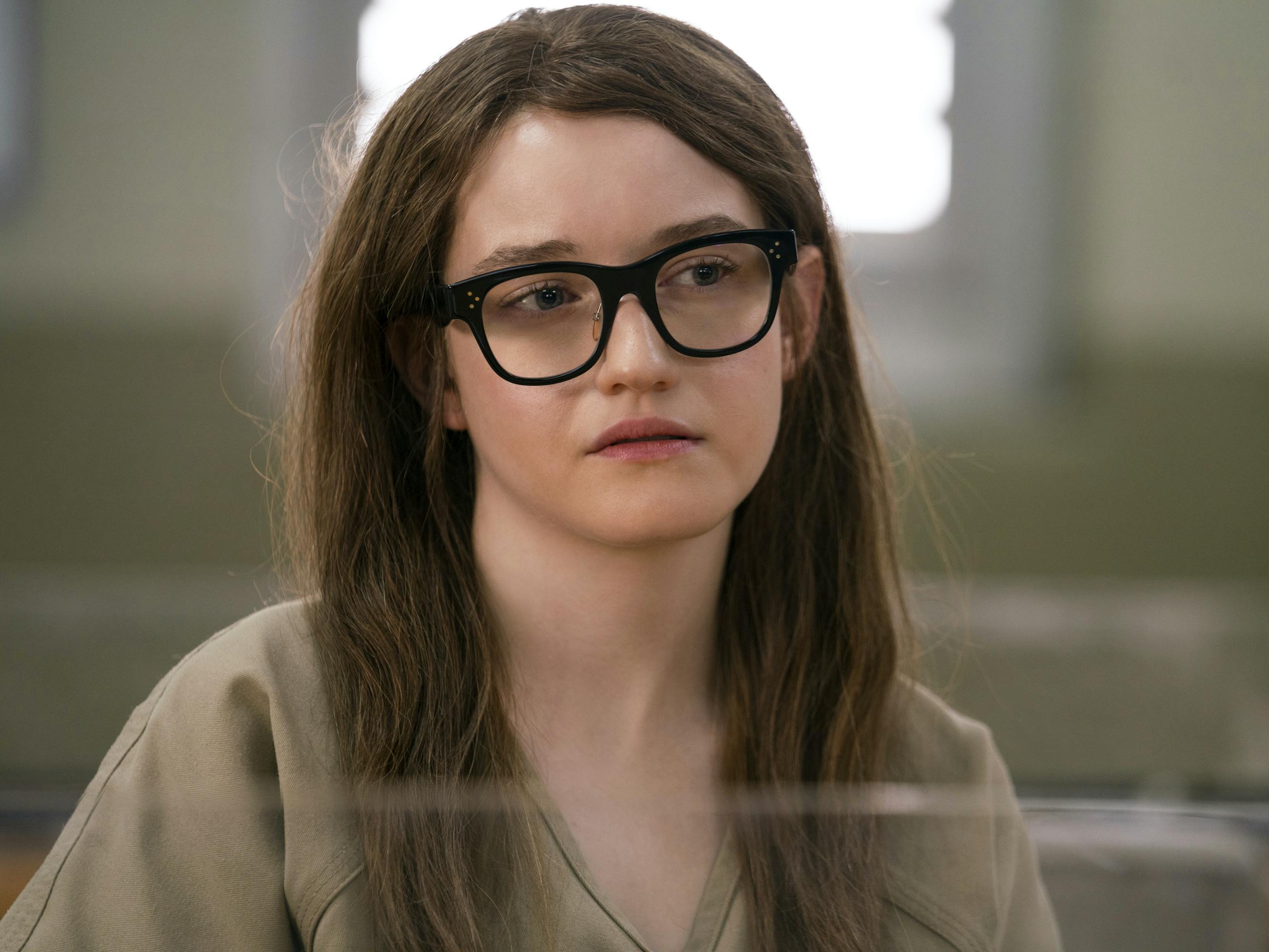 Anna Delvey (Julia Garner) wears dark-rimmed glasses and a beige shirt.