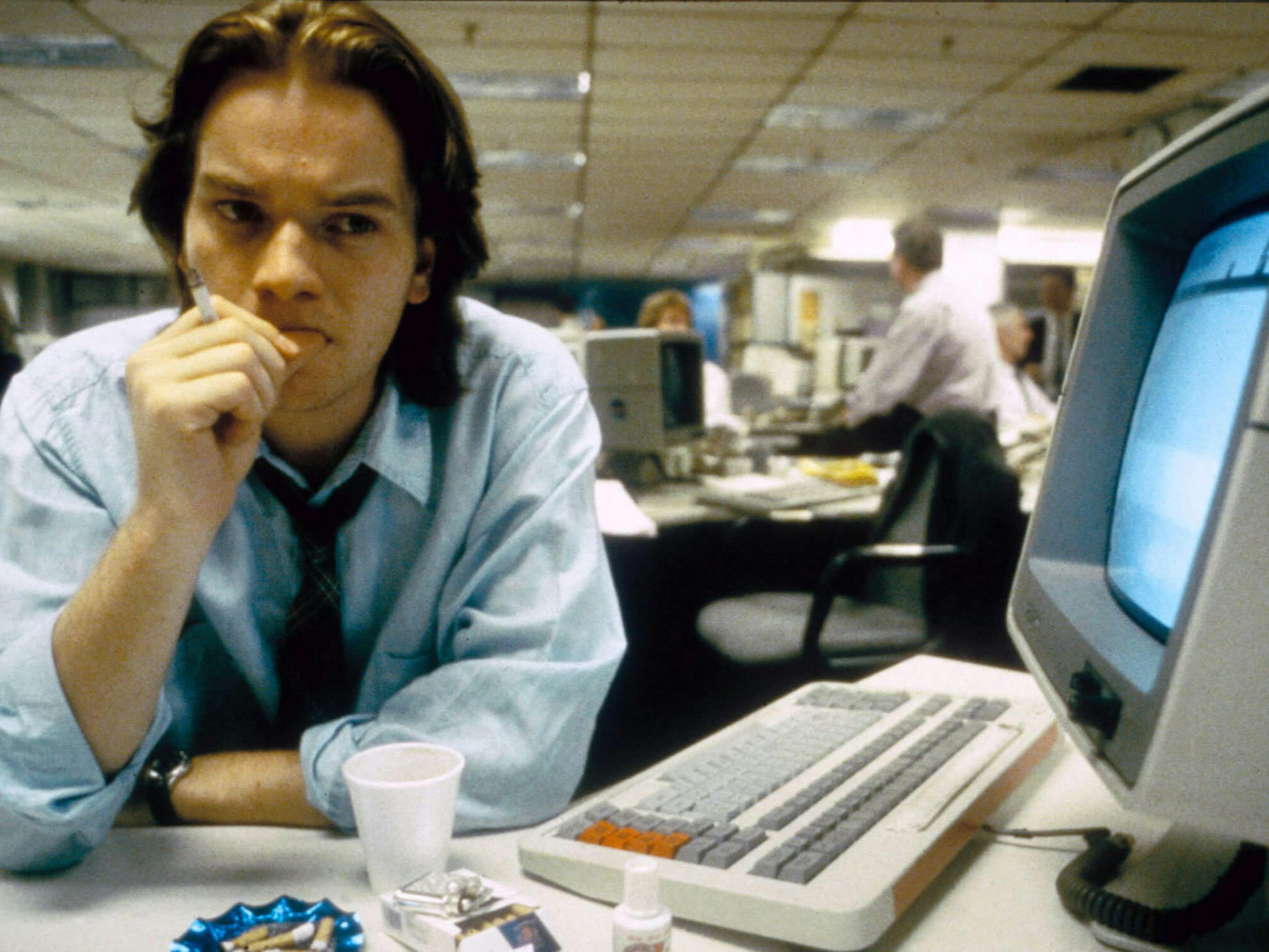 Alex Law (Ewan McGregor) in Shallow Grave sits at a desktop computer and keyboard. He wears a blue buttoned down and dark tie, and looks consternated.