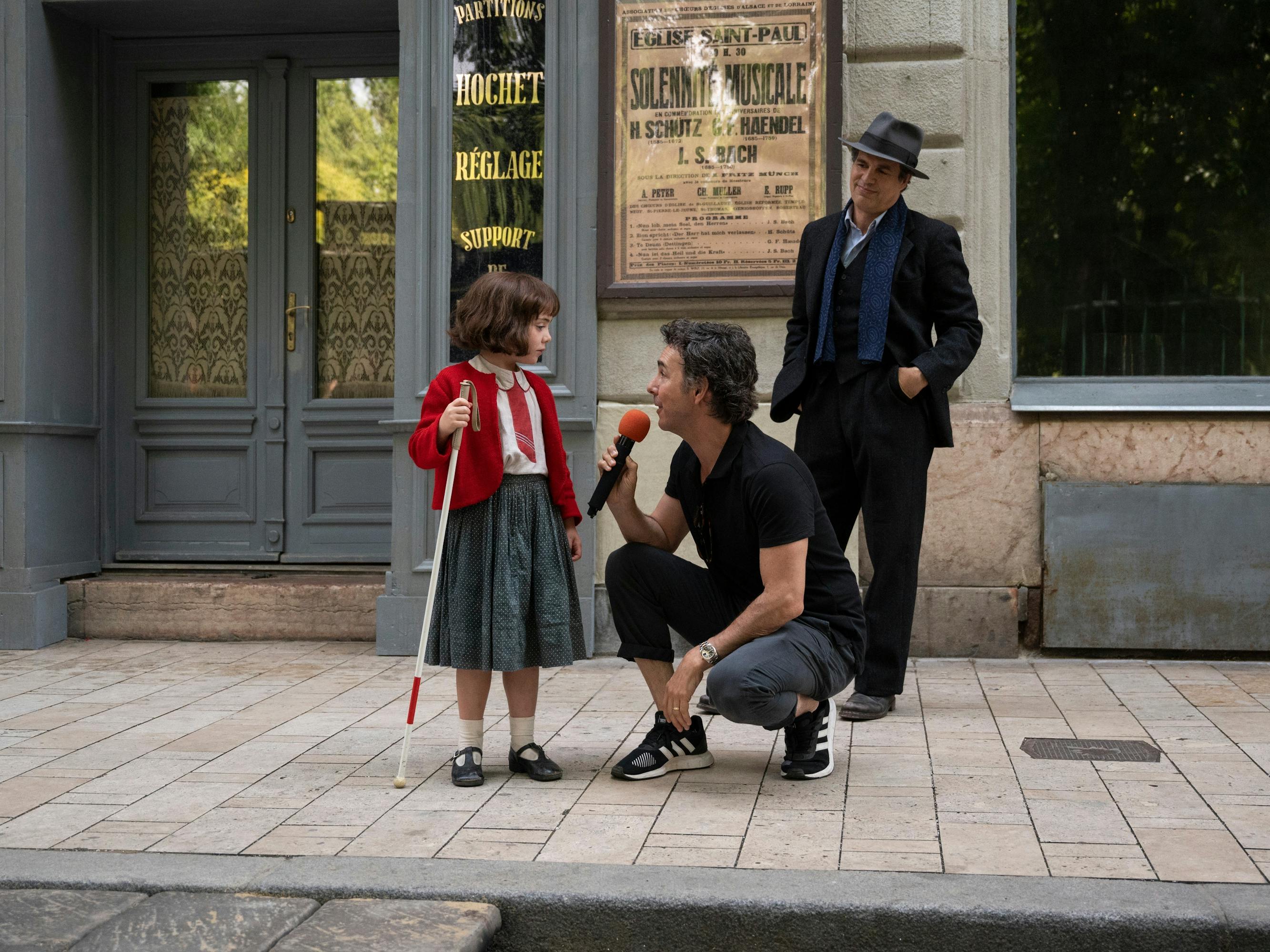 Nell Sutton, Shawn Levy, and Mark Ruffalo behind the scenes on set. Sutton looks downright precious in a long skirt, red cardigan, matching red tie, black mary janes, and holding a white cane. Levy holds a red mic up. Ruffalo stands behind the duo wearing an impressive hat and a blue scarf.