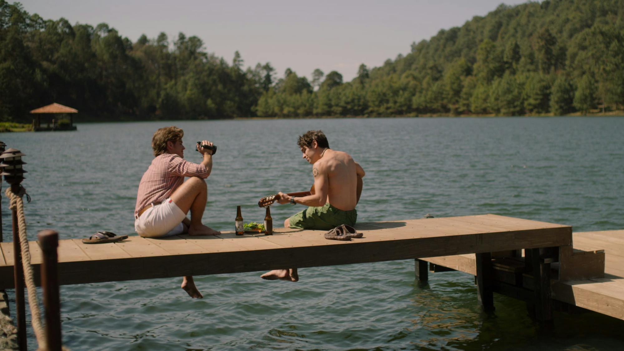 Two men sit on a dock. Water sloshes beneath their dangling feet as one man films the other playing guitar. Beside the men are two sets of flip flops and beer bottles. The lake and mountain behind them make the scene idyllic.