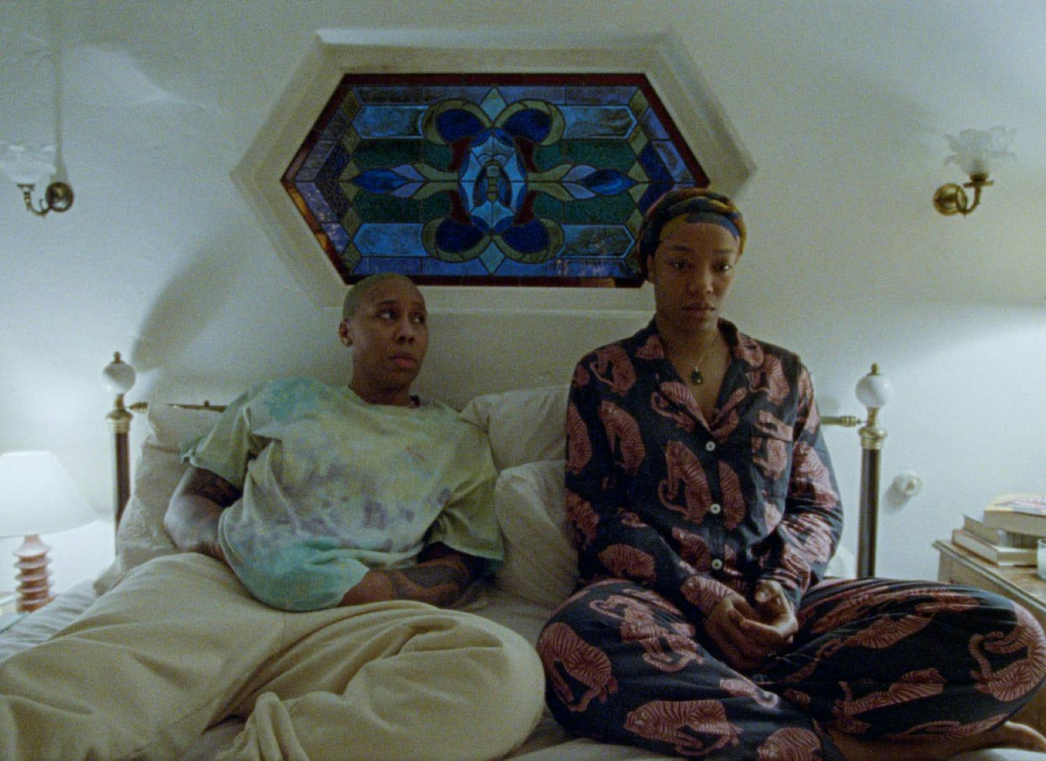 Denise (Lena Waithe) and Alicia (Naomi Ackie) sit in bed. Denise looks at Alicia with a concerned expression, and Alicia stares into space in very chic pajamas. On the wall is a stained glass window with blue, green, yellow, and red panes, as well as small lamps.