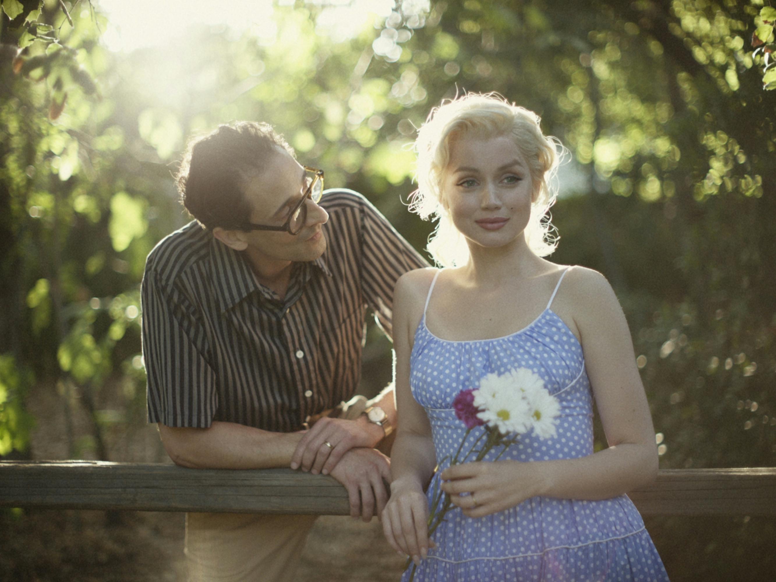 The Playwright (Adrien Brody) and Marilyn Monroe (Ana de Armas) stand together at a fence. Brody wears glasses and a short sleeved shirt. Marilyn wears a blue dress and carries a bouquet of flowers. 