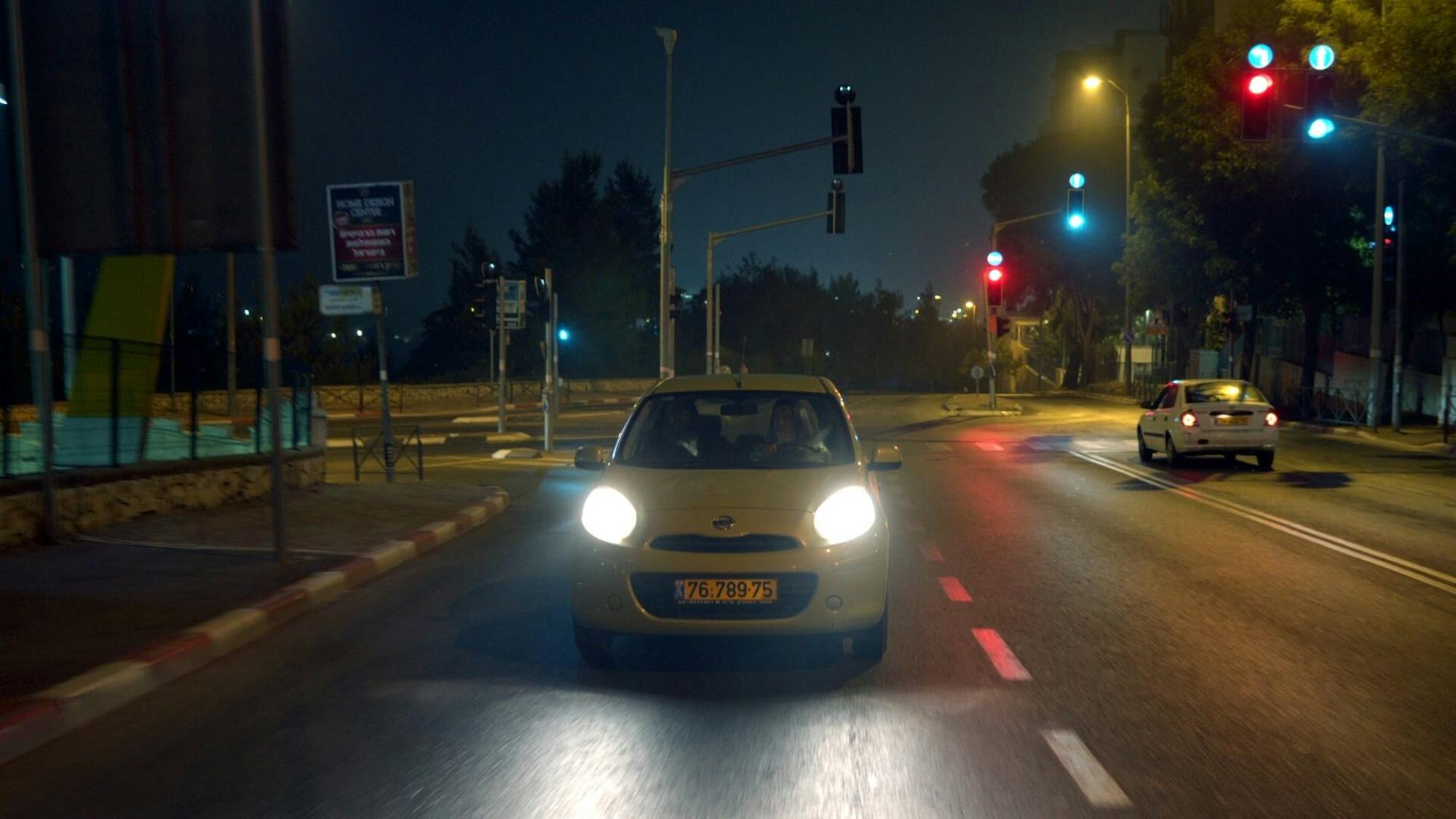 A shot of a white car on a street. The headlights are on. It is nighttime and there is one other car on this empty street.