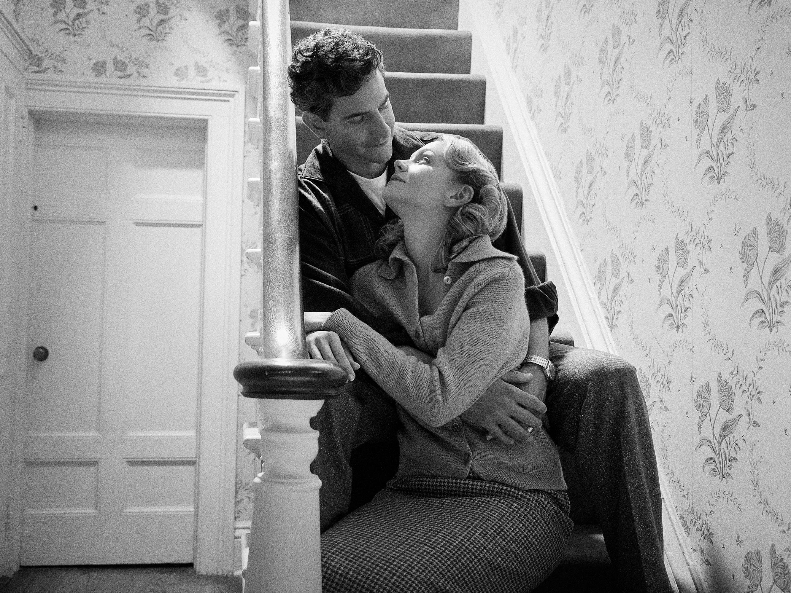 Leonard Bernstein (Bradley Cooper) and Felicia Montealegre Cohn Bernstein (Carey Mulligan) sit on the stairs together. The walls behind them are painted with patterned wallpaper. 