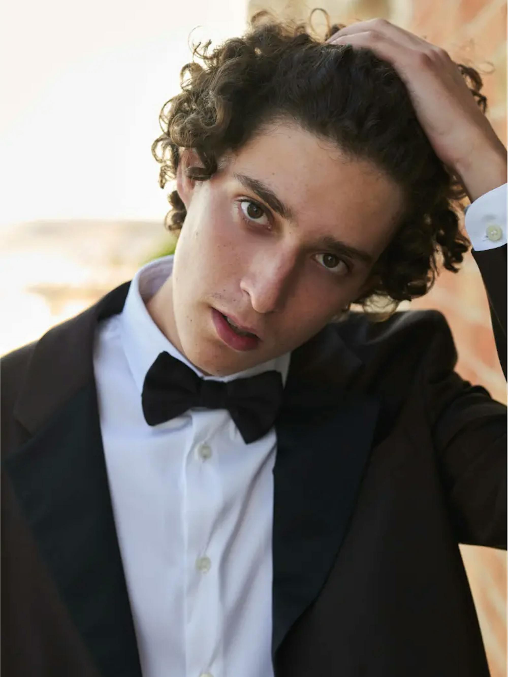 Filippo Scotti wears a tux and bowtie as he brushes his curls off his face in this close up shot.