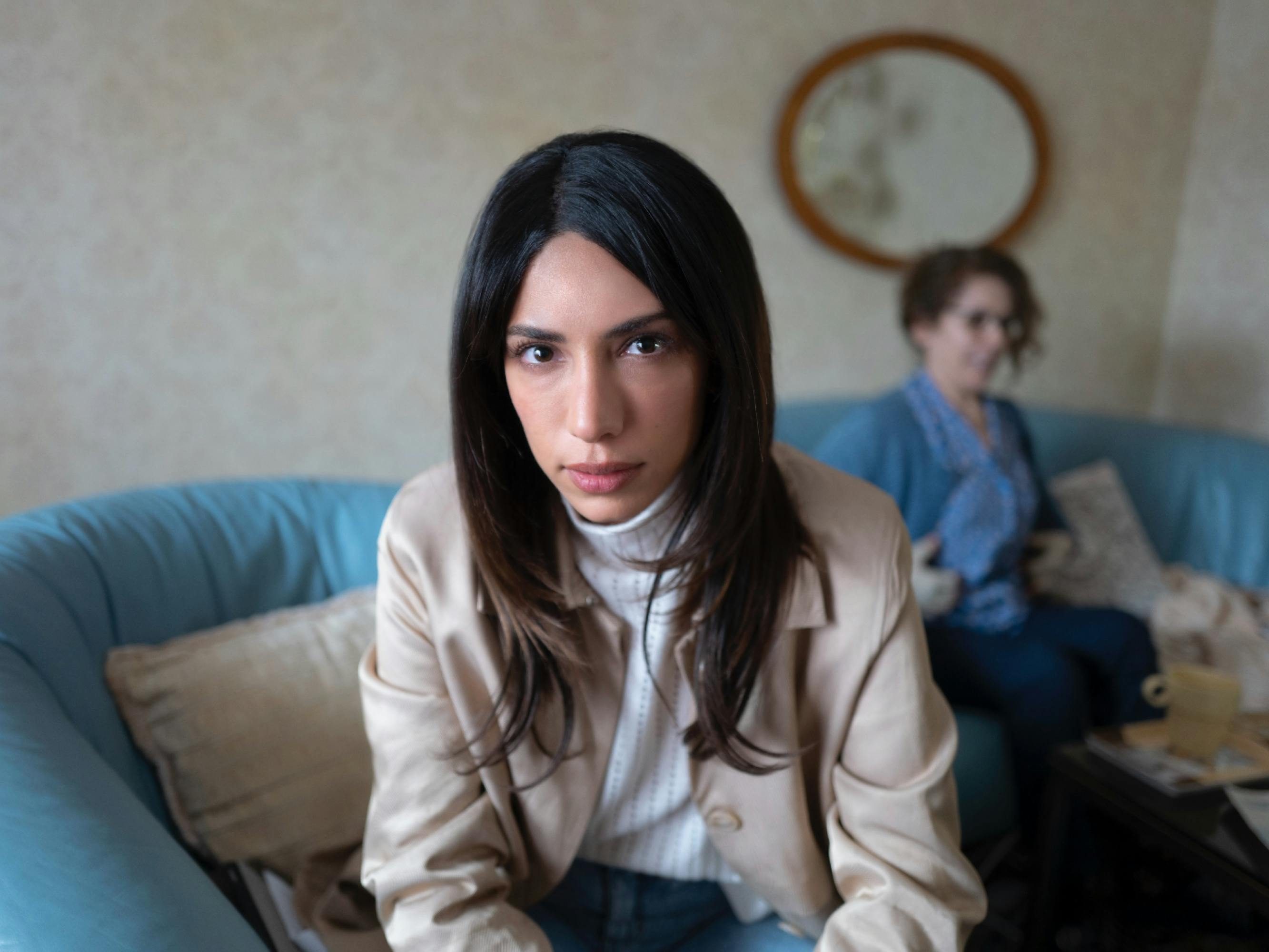Evin Ahmad, who plays Leya, wears a white turtleneck, tan coat, and looks directly at the camera. In the background there is a woman sitting on a turquoise couch.