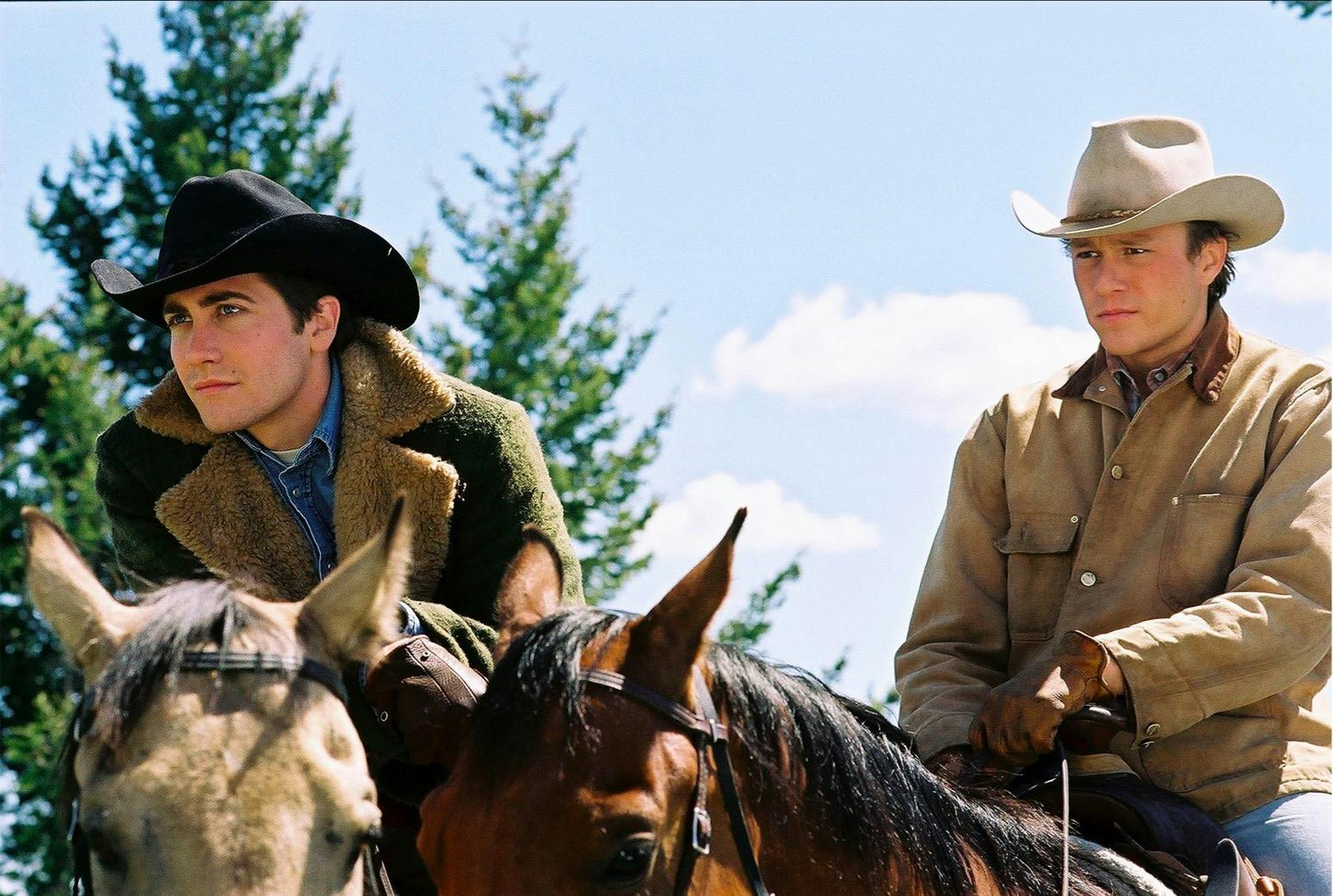 Jack Twist (Jake Gyllenhaal) and Ennis Del Mar (Heath Ledger) in Brokeback Mountain (2005) sit on horses side-by-side. Gyllenhaal wears a fur coat, dark hat, and the most epic denim shirt in all of film history. Ledger wears a tan jacket and tan hat. The sky is blue and there are green trees in the background.