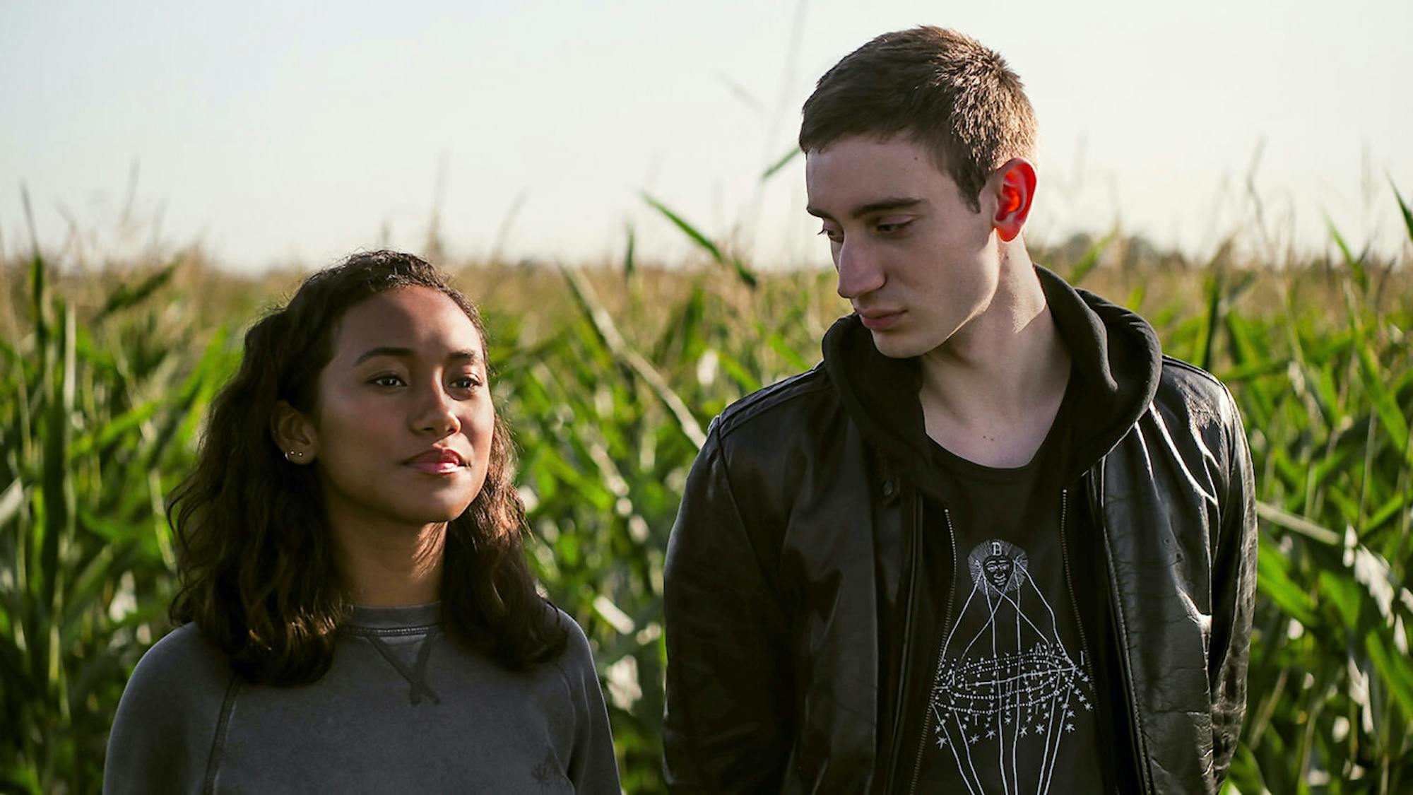 Makani Young (Sydney Park) and Ollie Larsson (Théodore Pellerin) stand in a corn field wearing dark clothing. Ollie looks to his right at Makani, while she looks off into the distance.